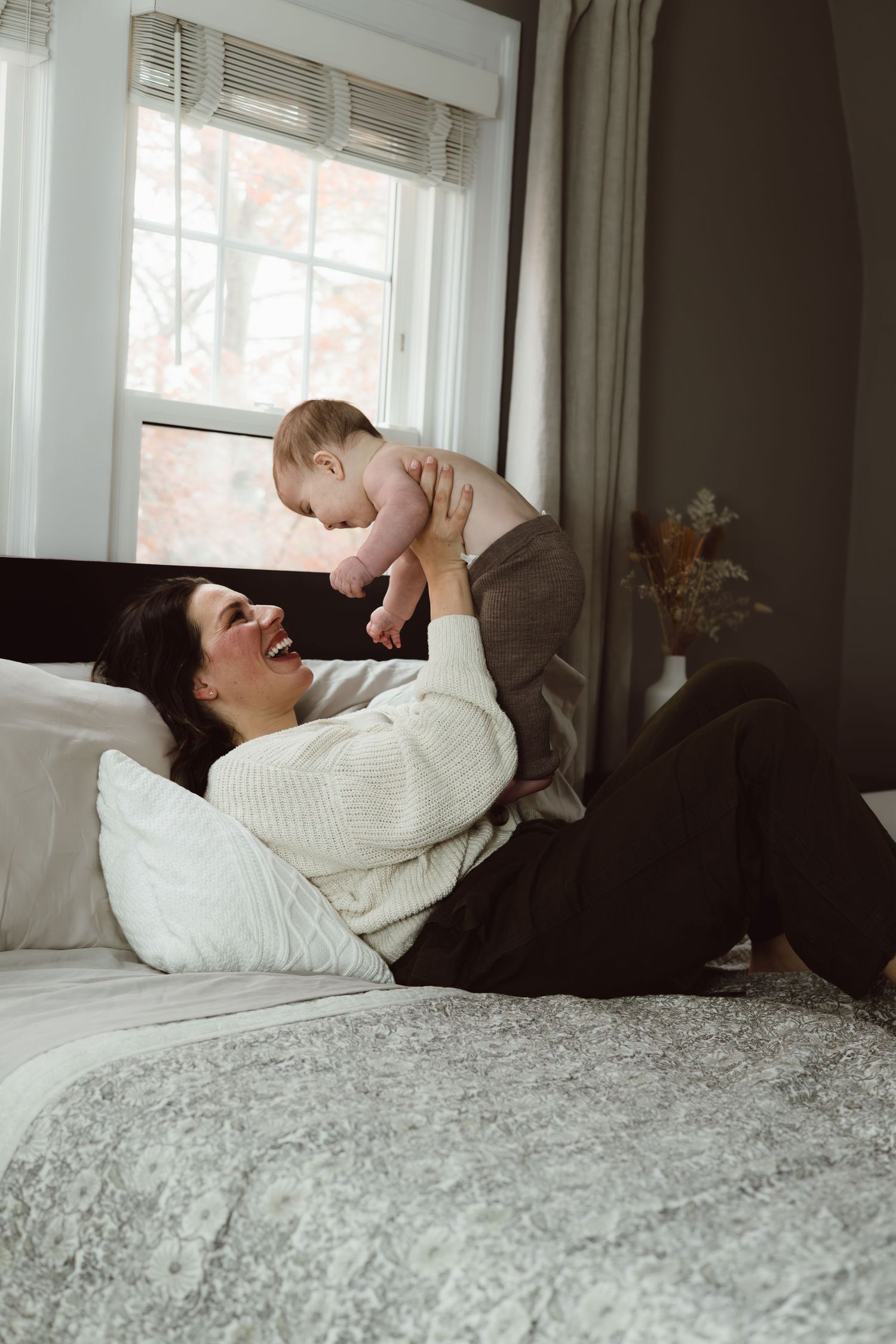 A mom in a white sweater relaxes on a bed with gray bedding while holding her 8 month old above her head laughing.