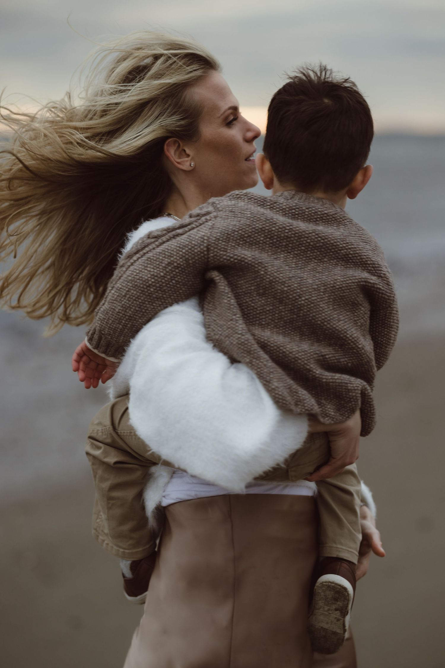 A mother and son on a beach with wind blowing through blonde hair as they share a tender moment at sunset.