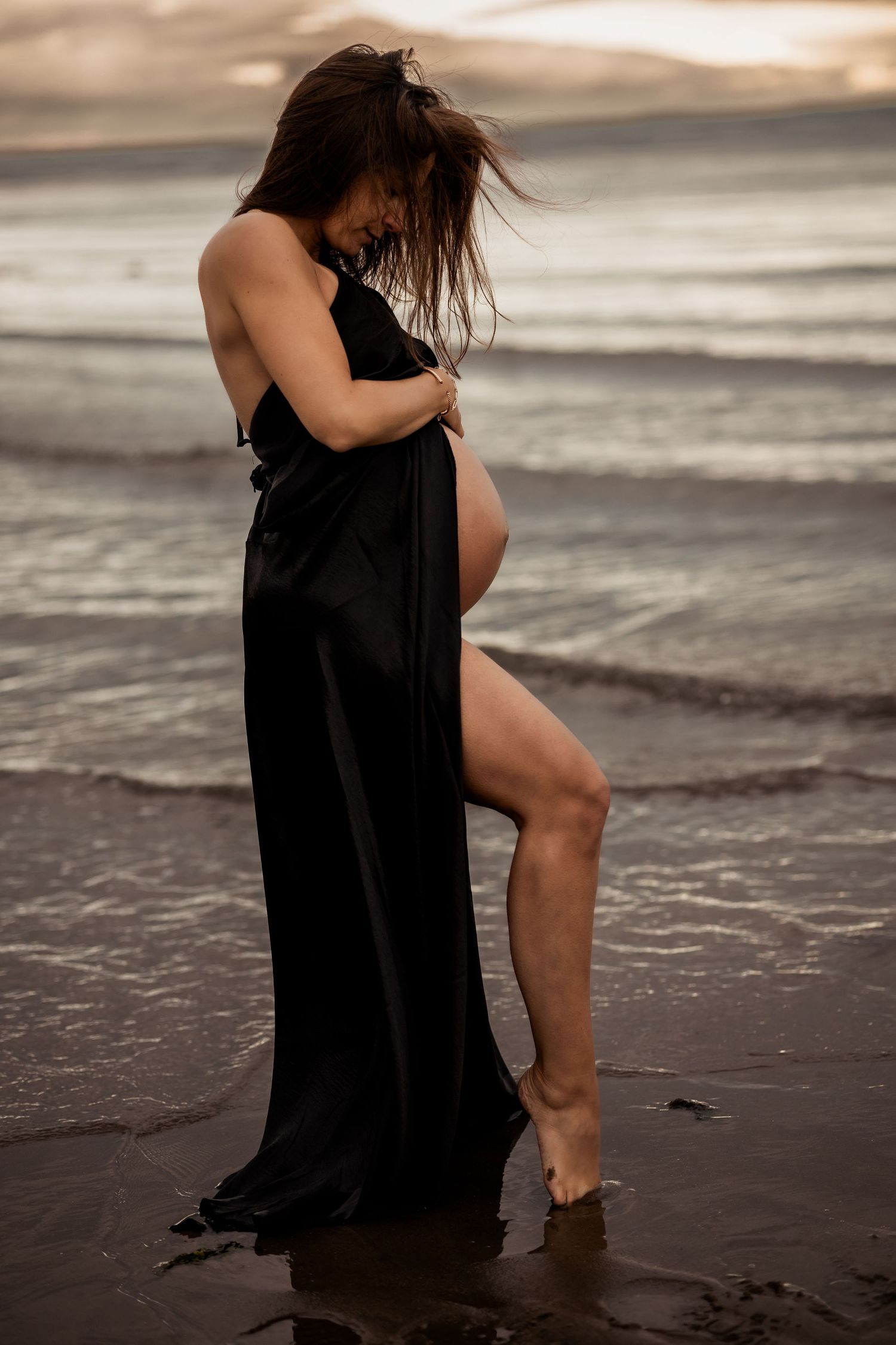 A pregnant woman in a flowing black dress poses artistically at the ocean's edge during sunset.