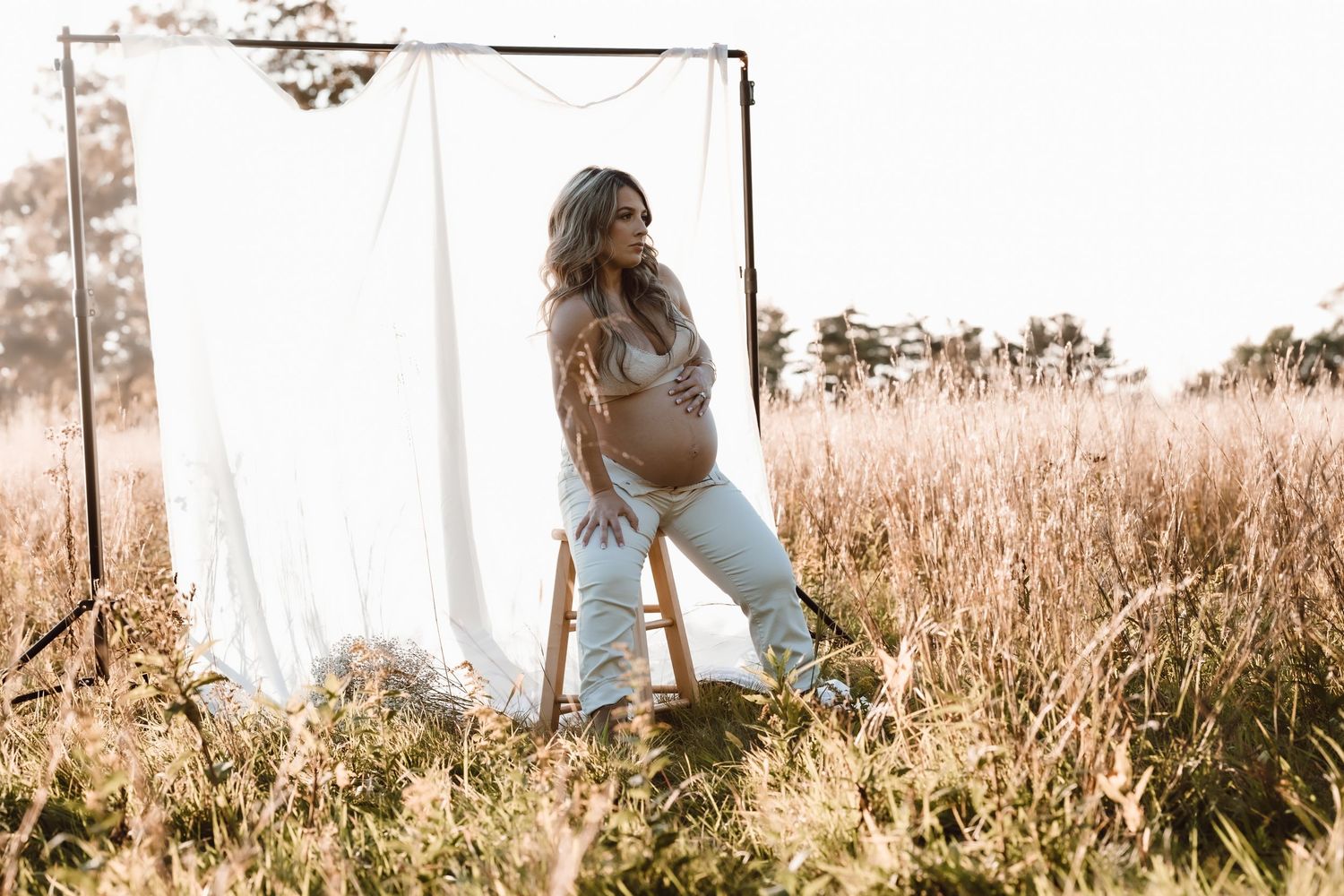 A pregnant woman stands in a sunlit field of tall golden grass during golden hour in front of white sheet backdrop sitting on a stool.