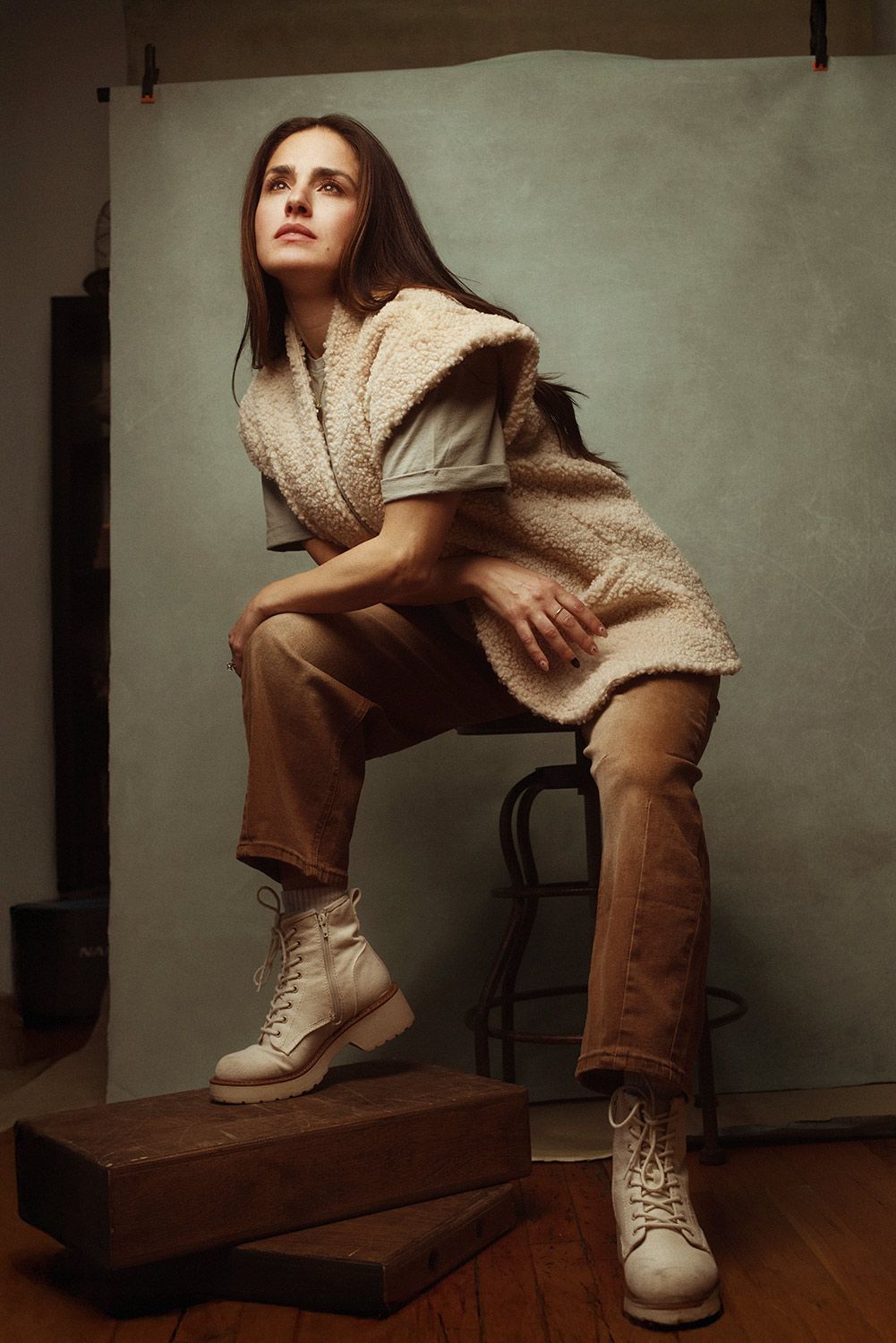 A motherhood photographer posting for a headshot showing white combat boots and brown pants on a wooden chair in moody lighting.