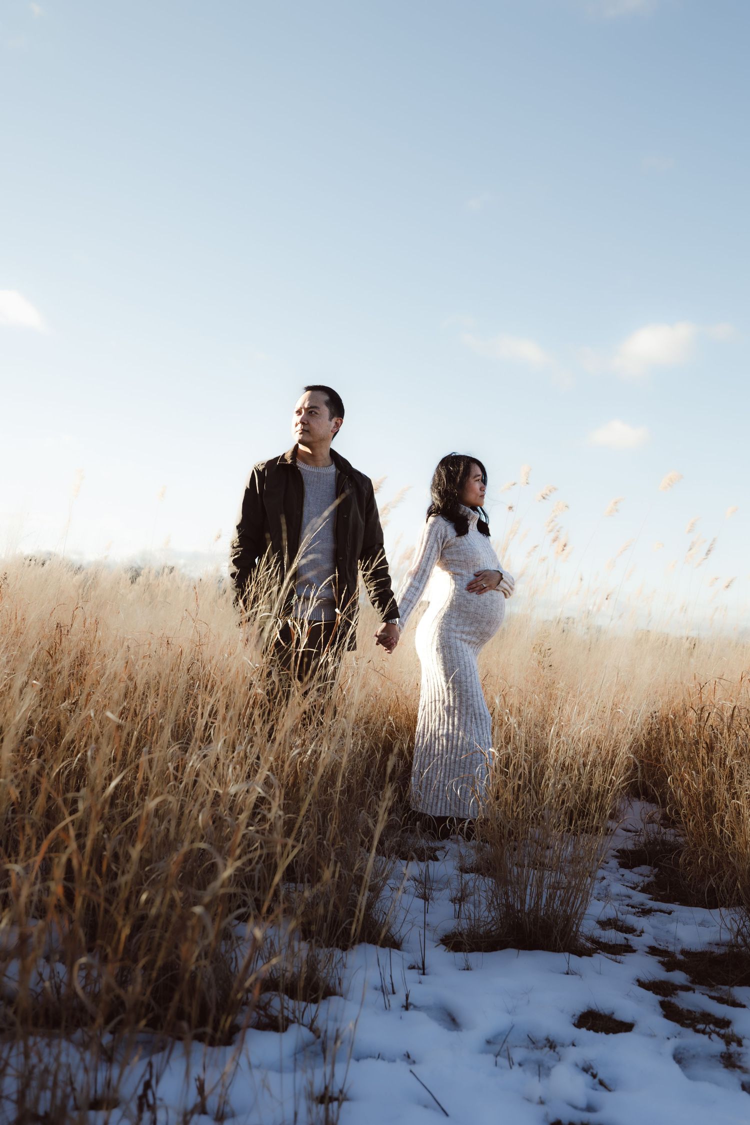 A couple during a maternity session walk together through a snowy winter field with tall dried grasses on a bright sunny day.