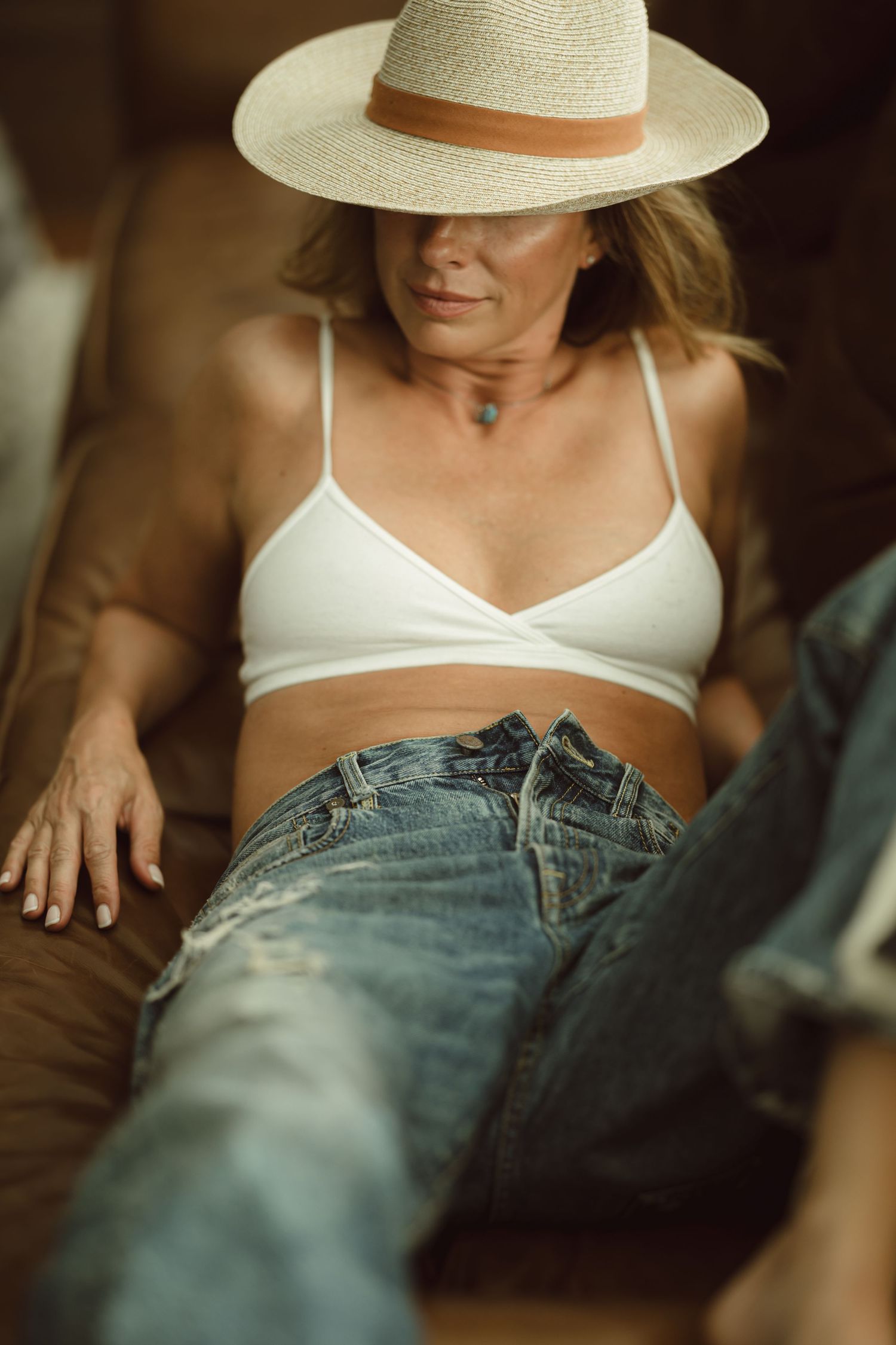 Woman wearing white cotton bra and jeans lounges on brown leather sofa while wearing a cream-colored sun hat.