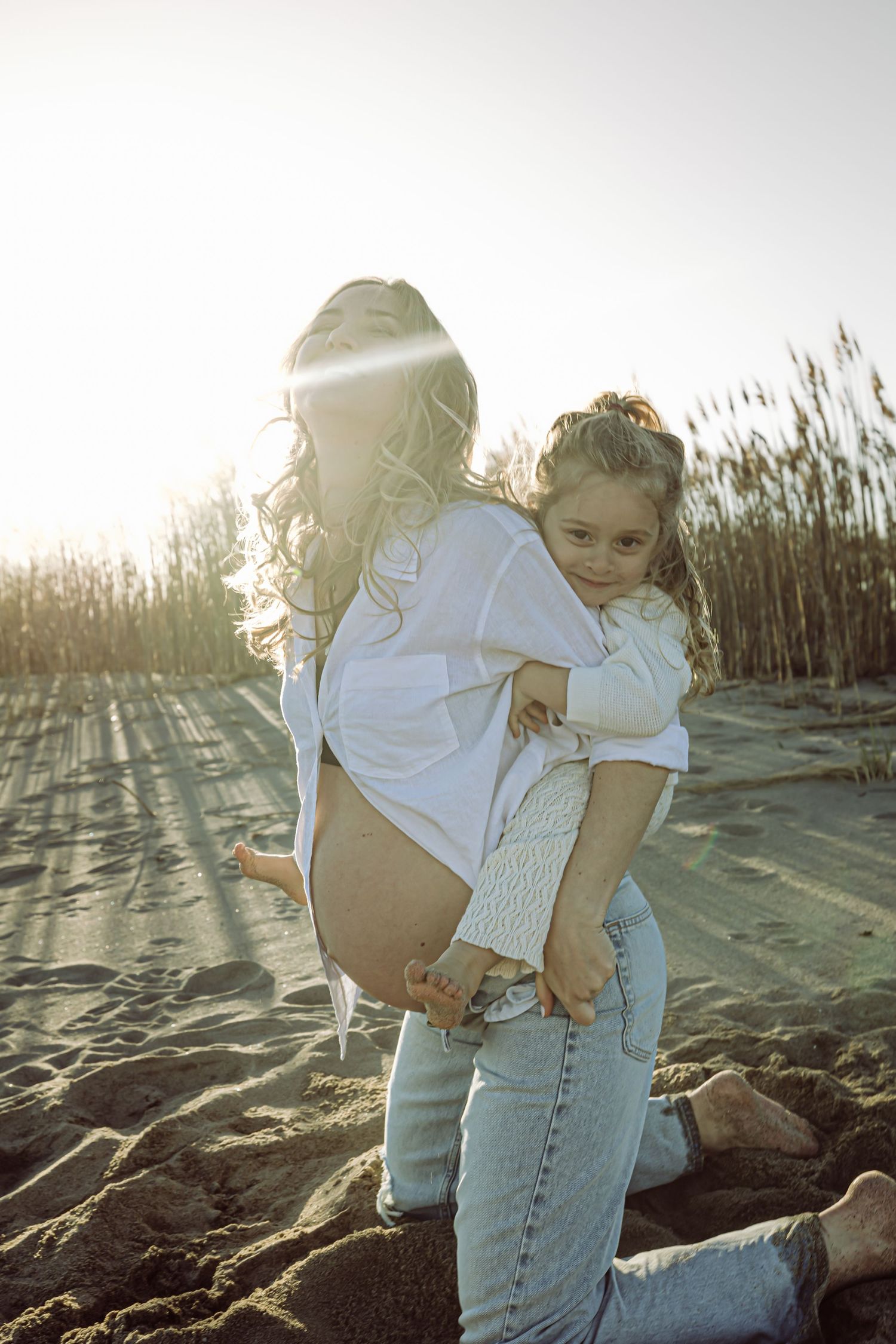A pregnant mother carries a child on her back on sandy beach at sunset with tall grass in background. Both laughing in joy during maternity.