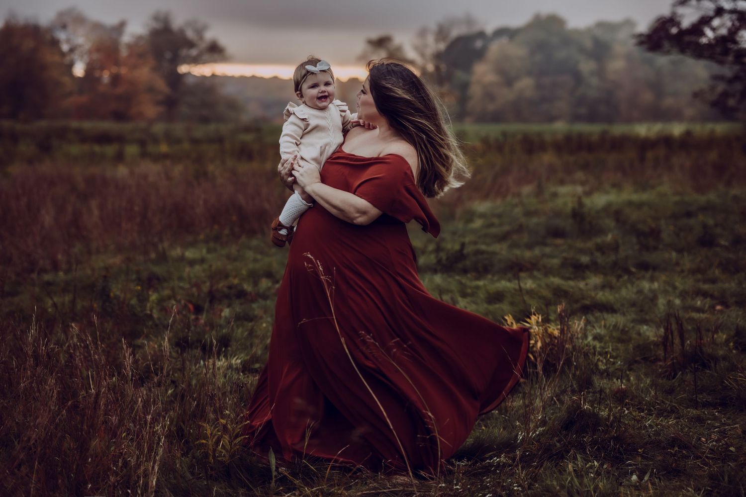 A mother in a flowing burgundy dress twirls gracefully in a misty autumn field at dusk holding her 8 month daughter for a family photoshoot.