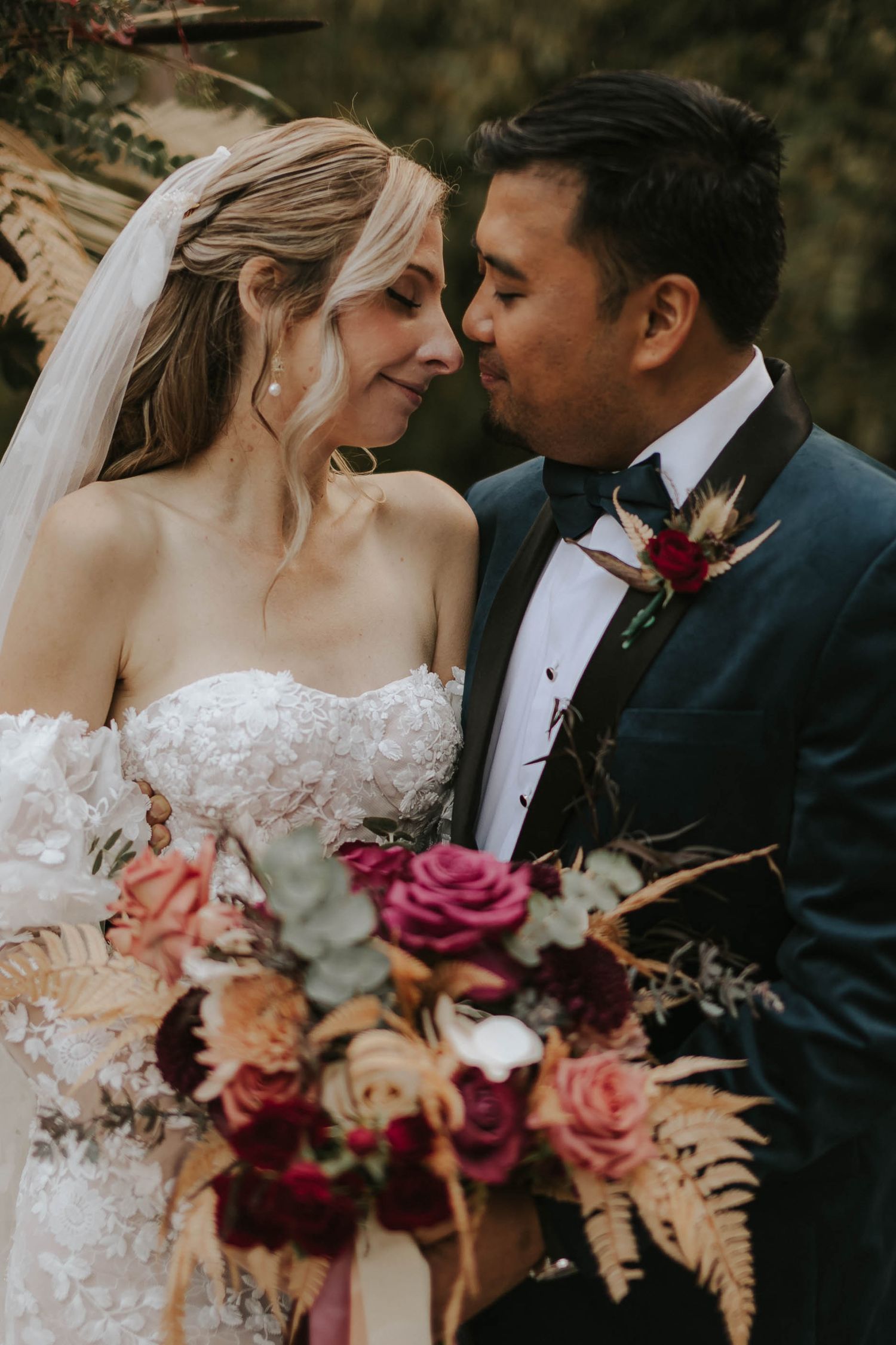 Close up of bride and groom sharing tender moment with burgundy and pink floral bouquet.