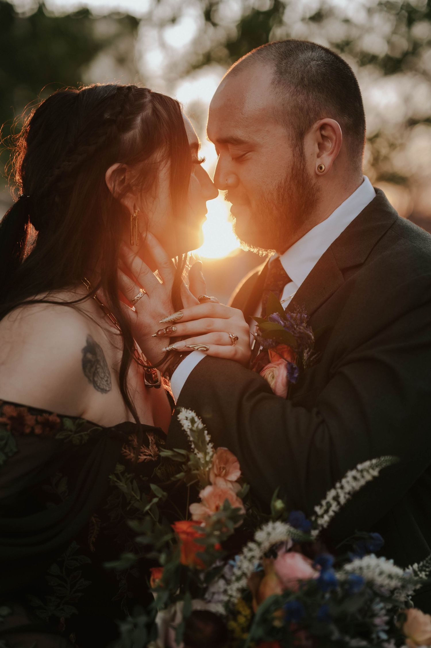 Couple sharing a romantic sunset moment, bride in black gown with tattoos, groom in dark suit, surrounded by colorful wedding florals.