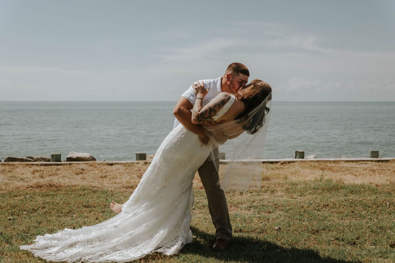Bride and groom share a romantic dip kiss on a grassy cliff overlooking the ocean on their wedding day.