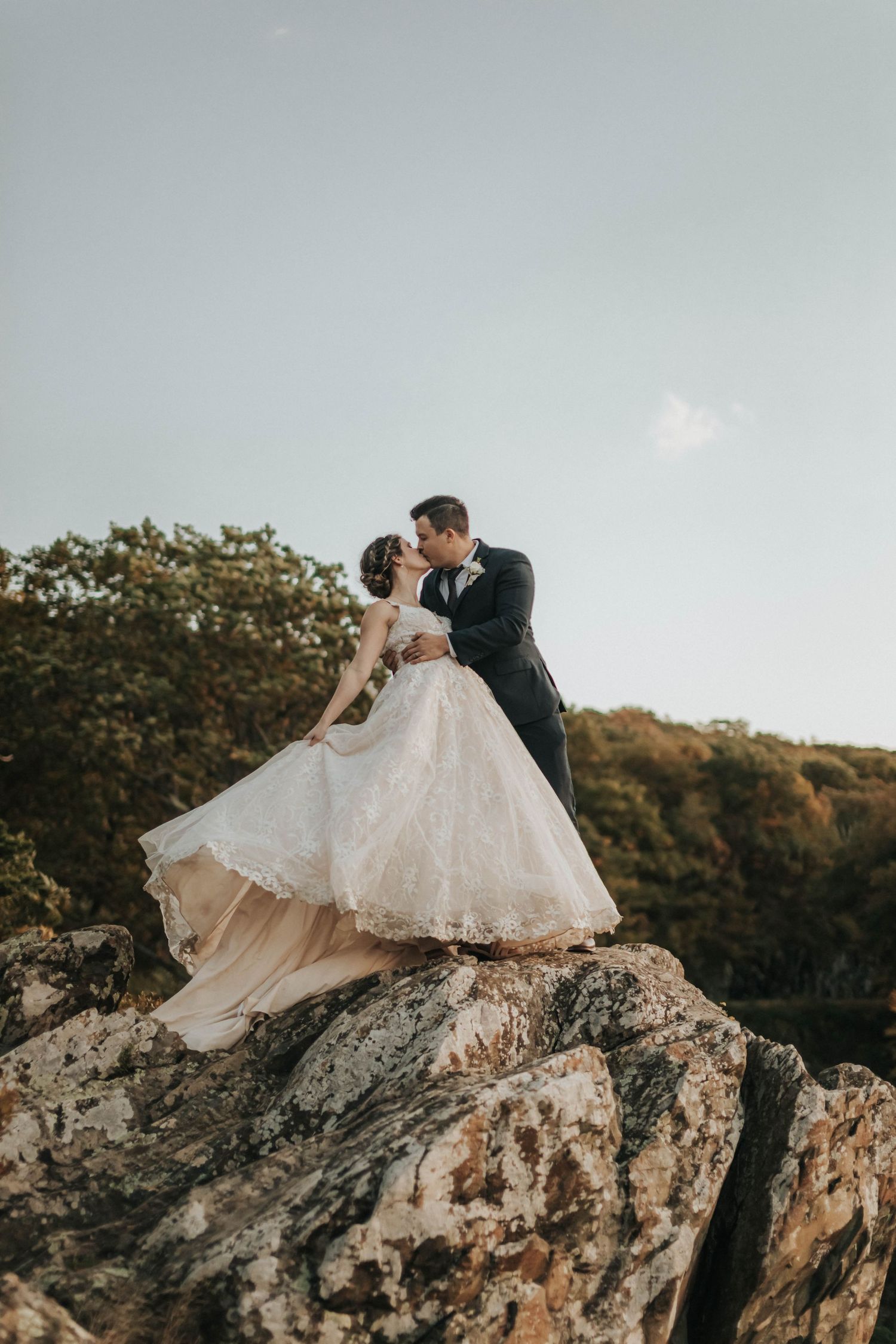 Newlyweds sharing intimate moment on rocky cliff edge in formal wedding attire.