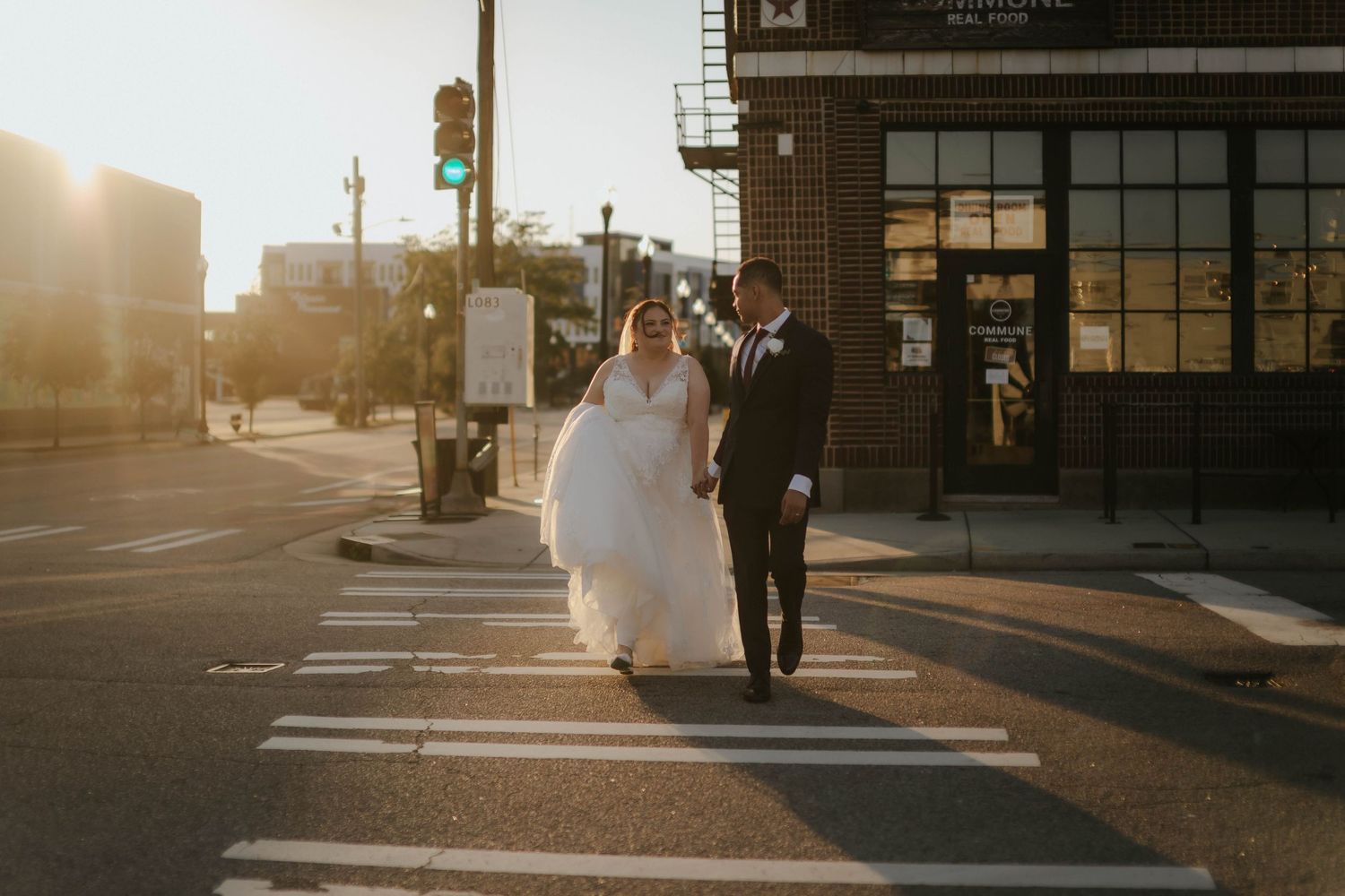Bride and groom crossing a sunlit urban street in wedding attire, golden hour light glowing behind them.