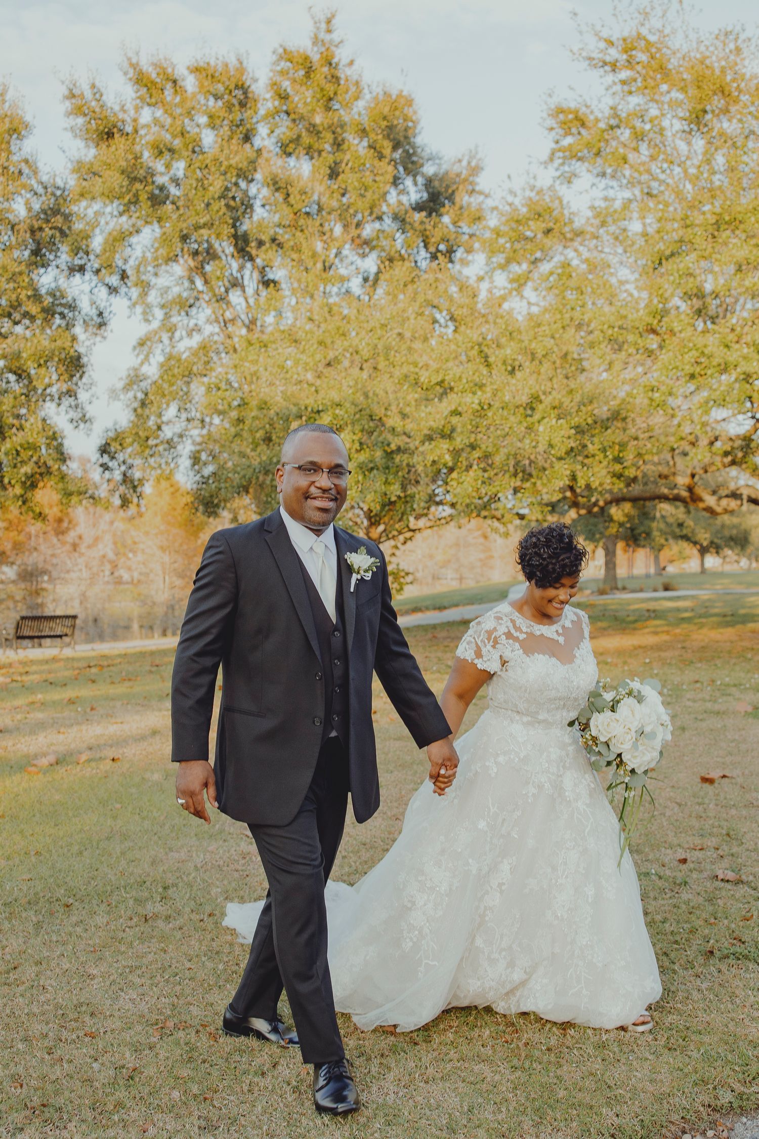 A bride and groom walk hand-in-hand through a park, she in lace gown, he in dark suit, with autumn trees behind.