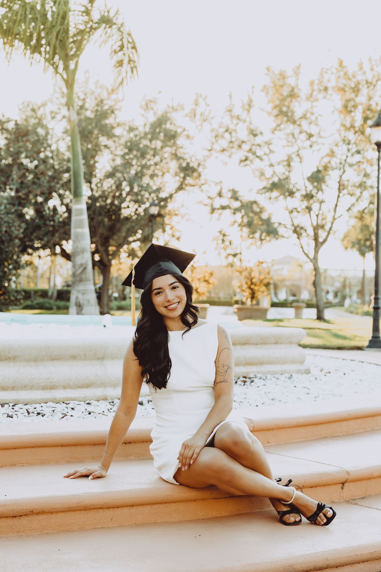 A woman in a white dress poses gracefully on a concrete ledge in a park setting during golden hour.