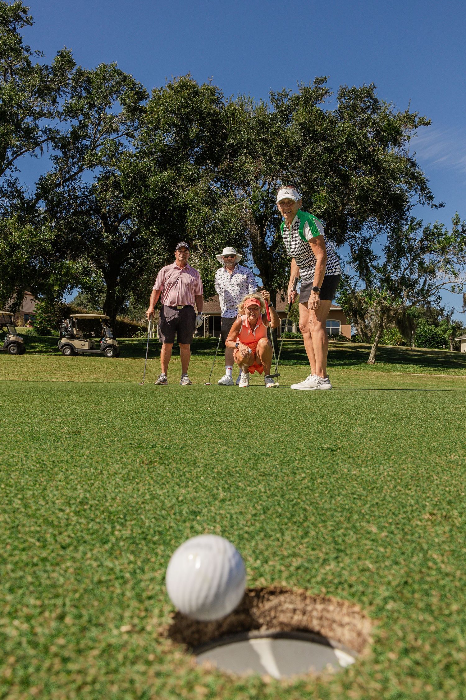 Golfers gather on a sunny green course with a golf ball in the foreground near a divot mark.