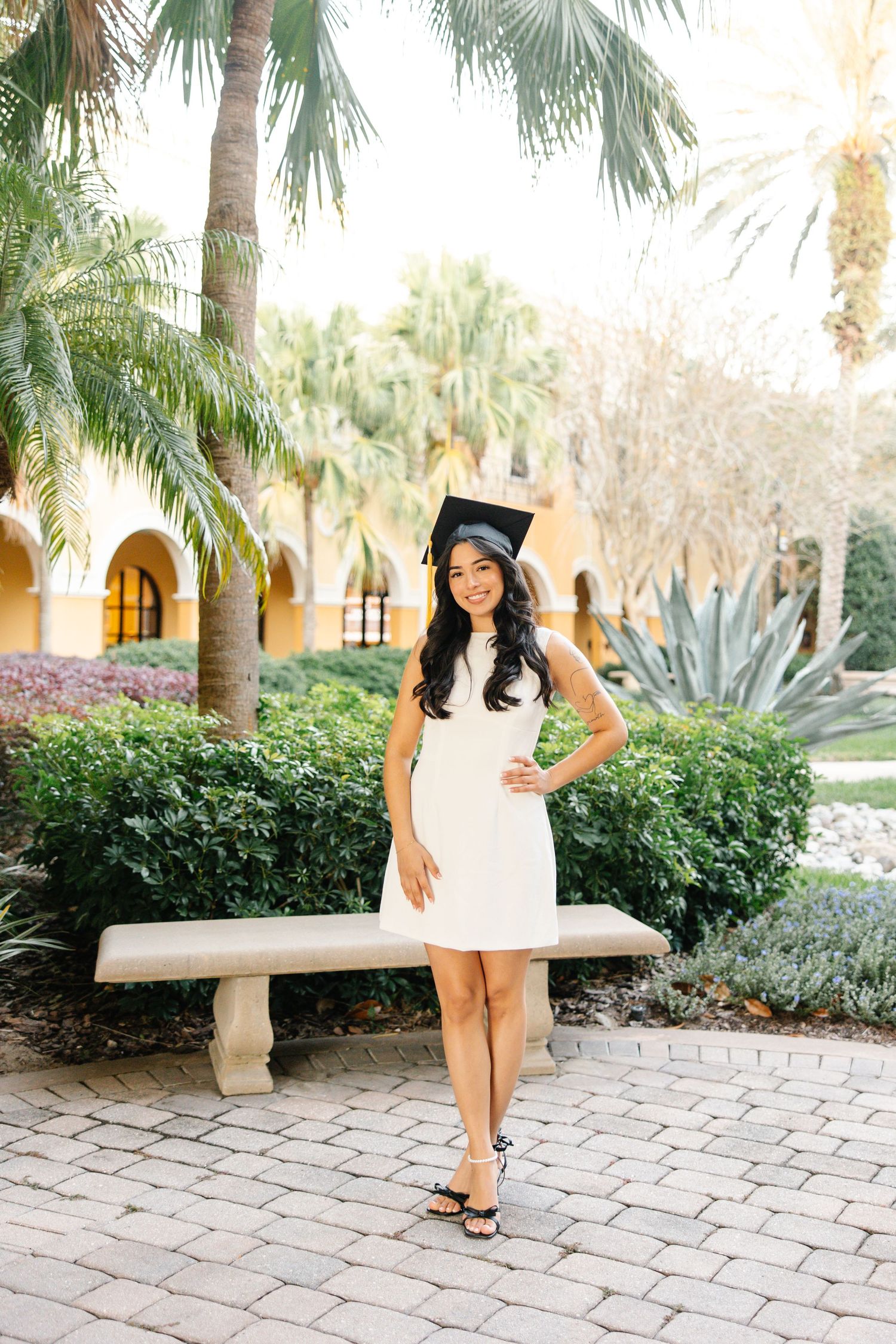 A graduate in a white dress poses with their diploma in a tropical garden setting with palm trees and stone pavers.