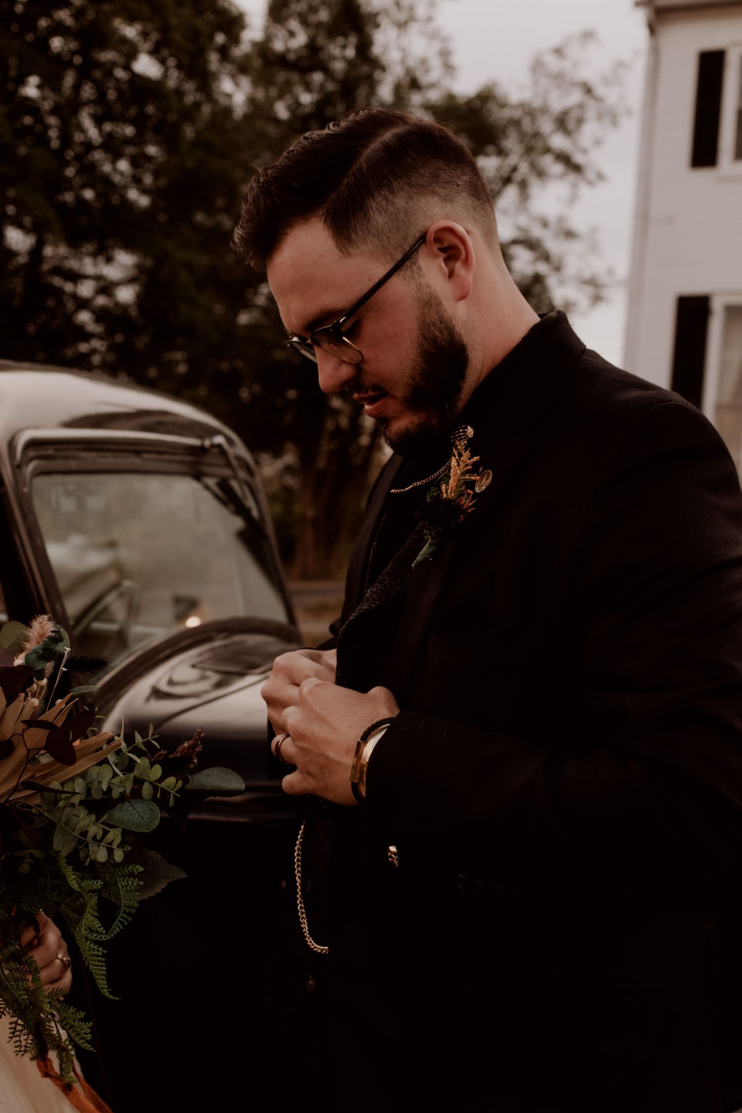 Person in black formal attire checking pocket watch outdoors.