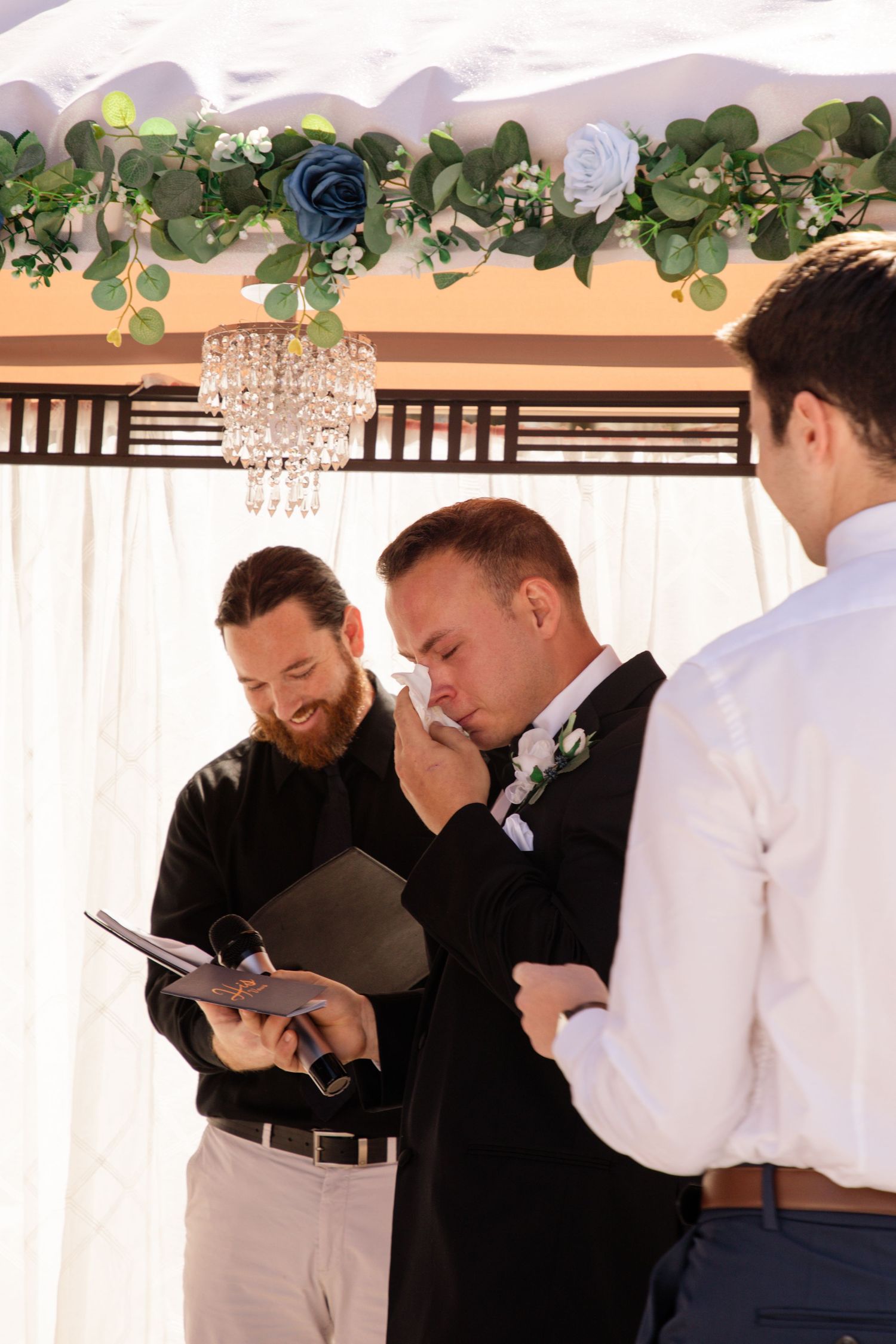 Emotional groom wiping tears during wedding ceremony while officiant reads vows under floral arch with chandelier.