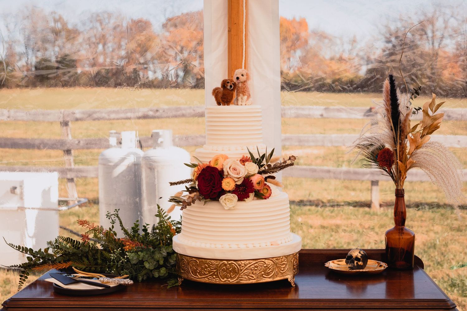Rustic wedding reception table with white cake, greenery, and autumn decor set against a golden field backdrop.
