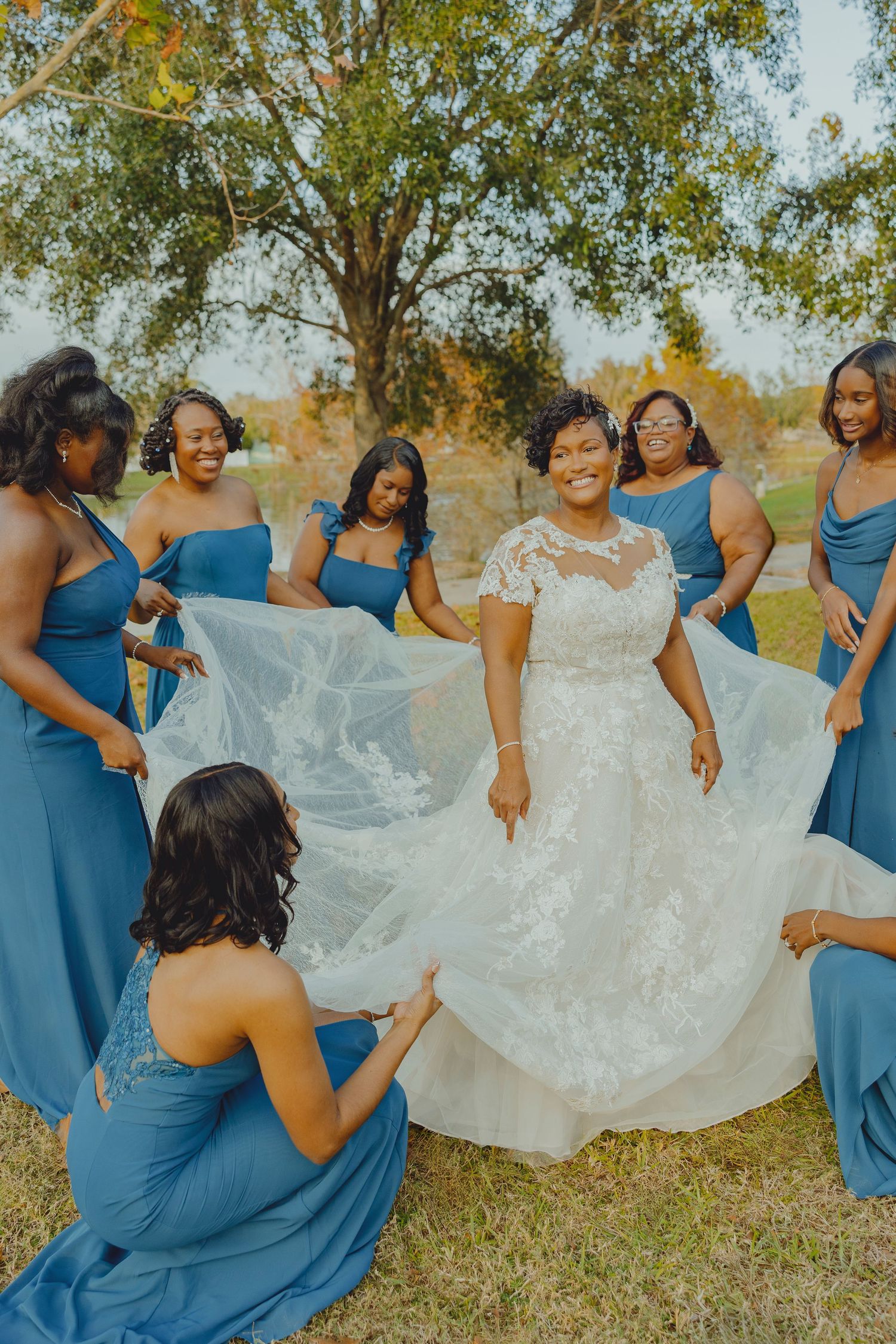 Bride in white lace gown surrounded by bridesmaids in blue dresses holding her veil outdoors.
