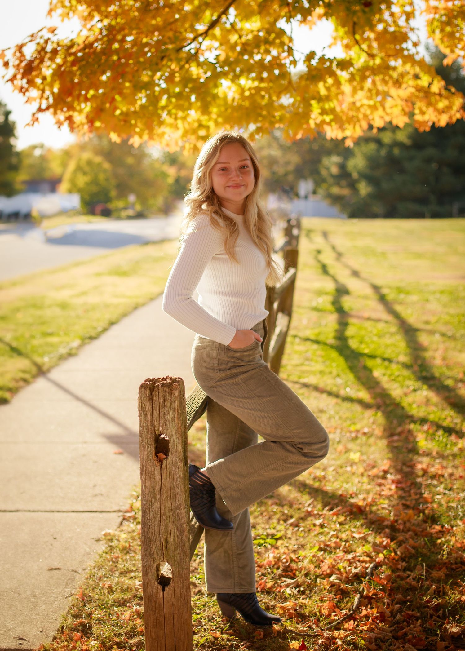 A person sits on a wooden post along a path during golden hour with autumn leaves scattered on the ground.