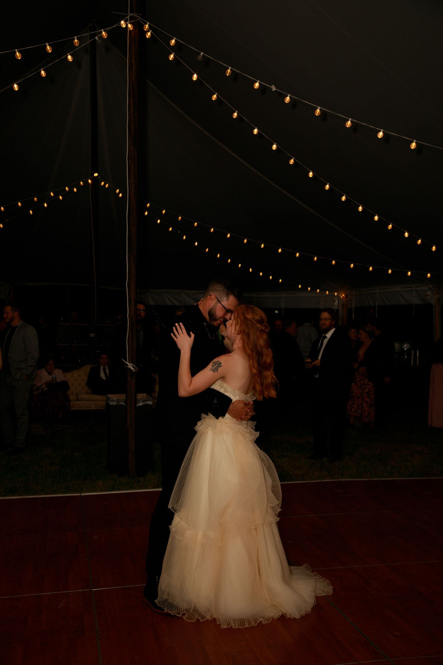 A couple dances together under twinkling string lights at an evening wedding reception.