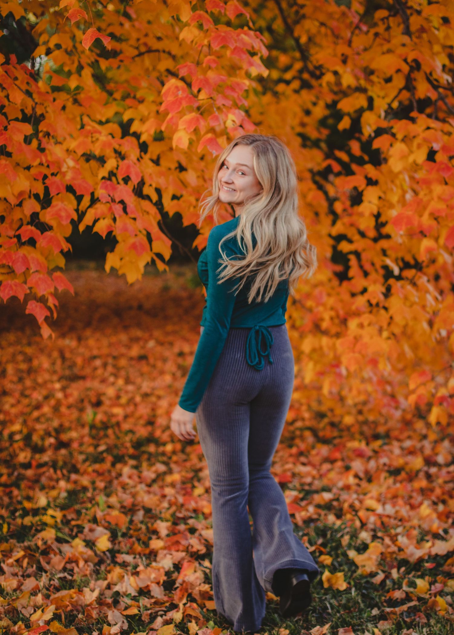 Person in teal sweater poses against vibrant orange autumn foliage in a scenic fall photoshoot series.
