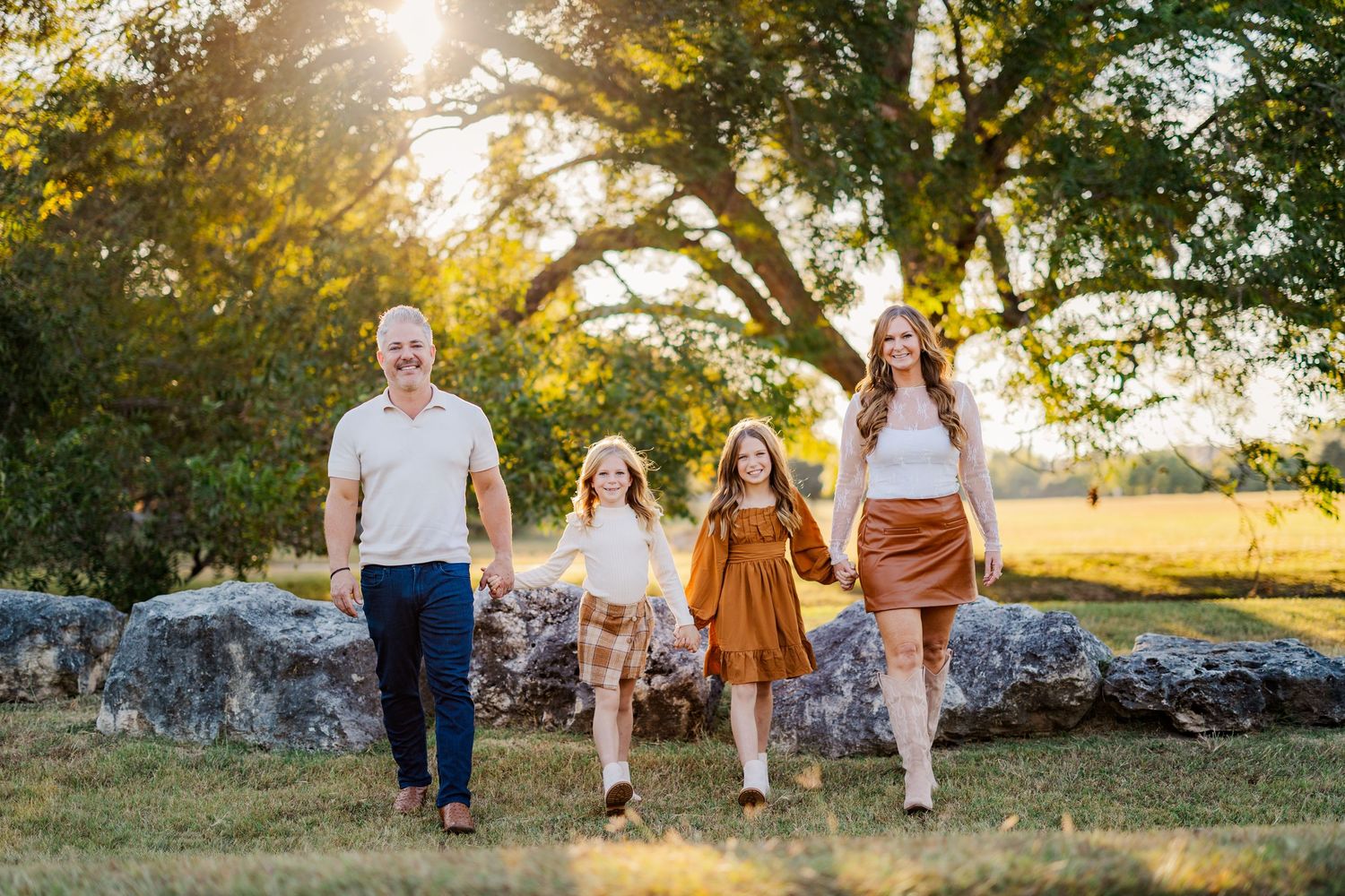 Family of four walking outdoors at golden hour near large rocks and trees in a sunny green field.