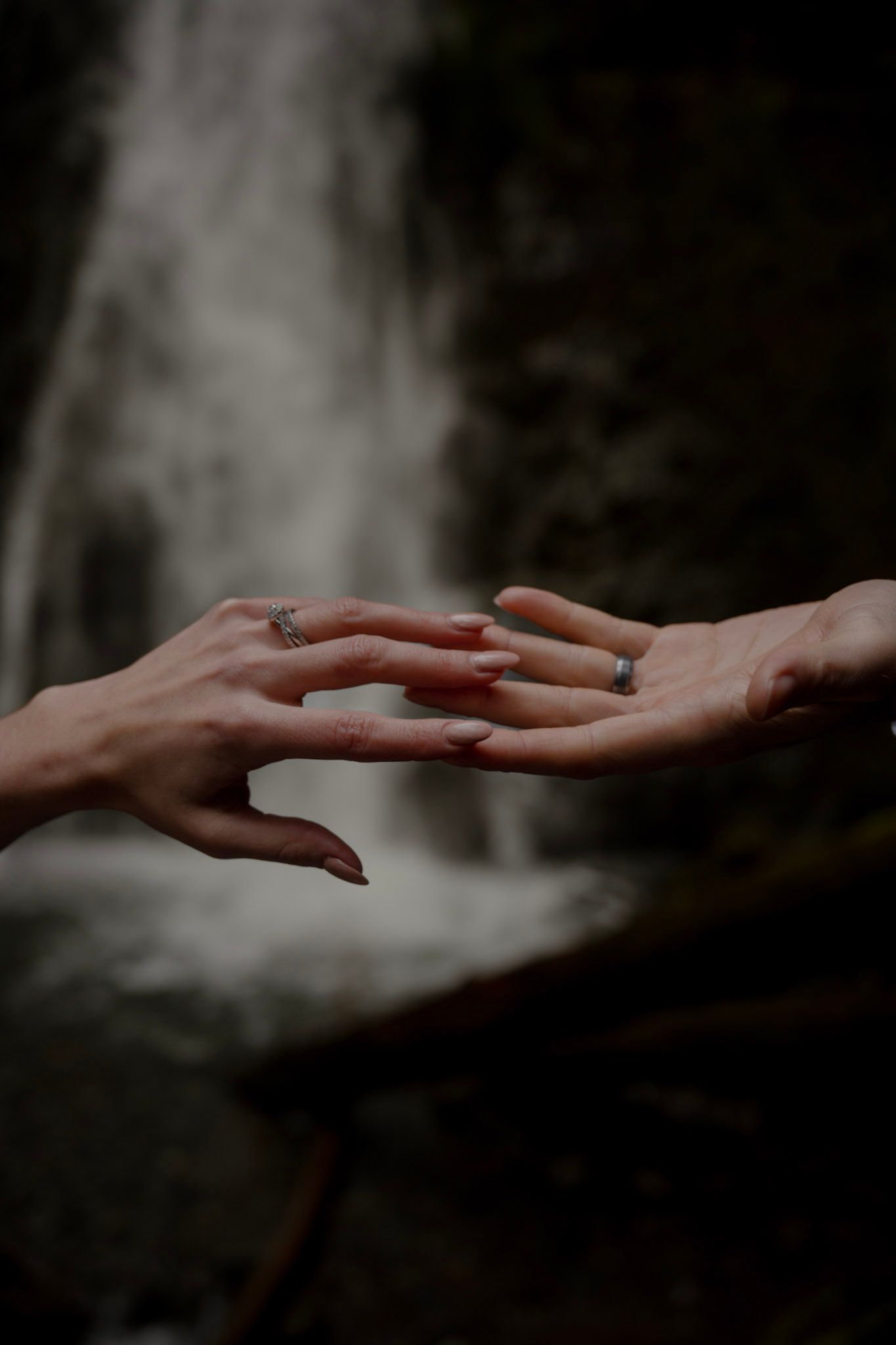 Two hands with wedding rings gently touch in front of a blurred waterfall background.