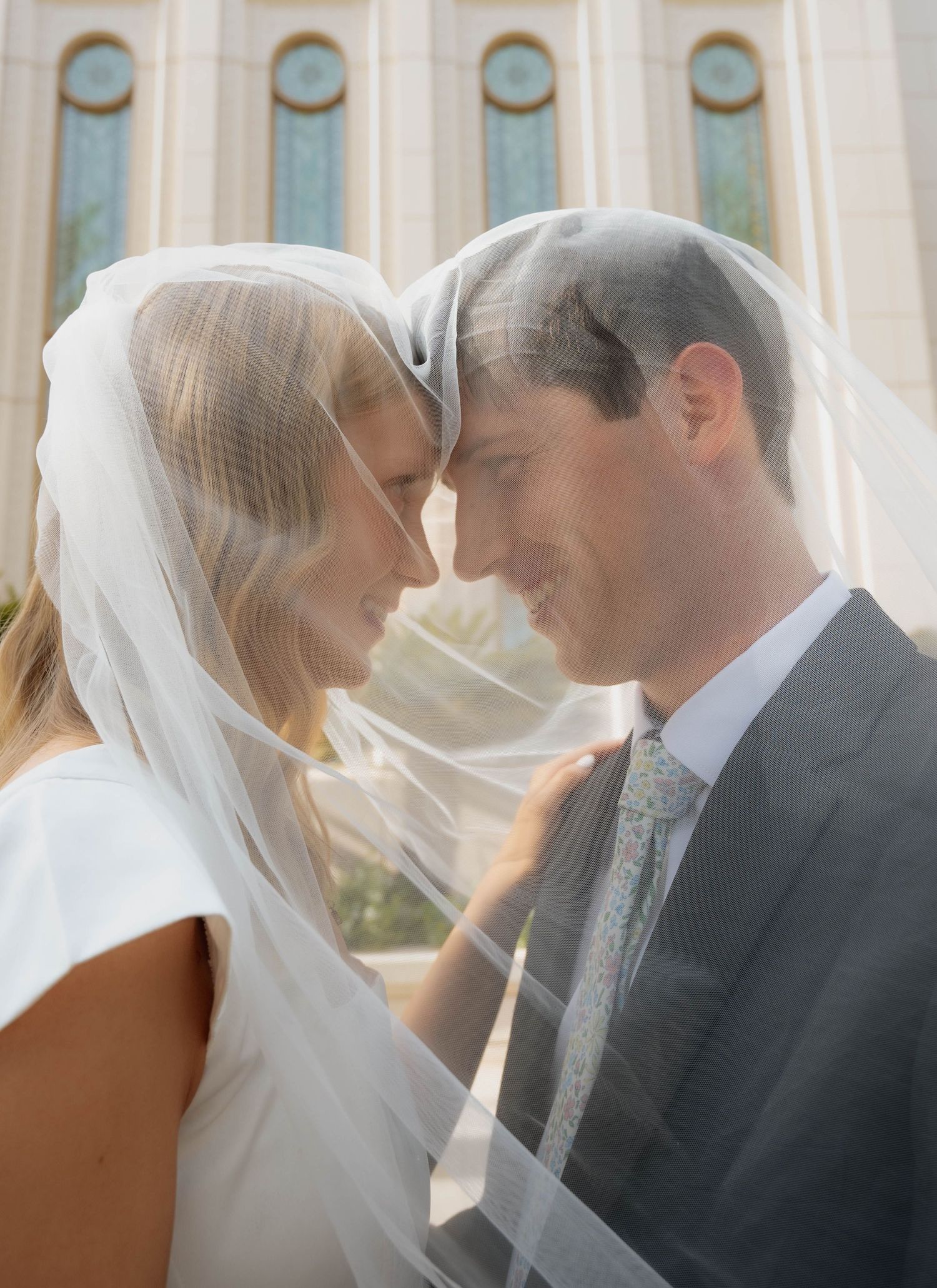 Newlywed couple shares intimate forehead touch beneath flowing veil outside temple on wedding day.