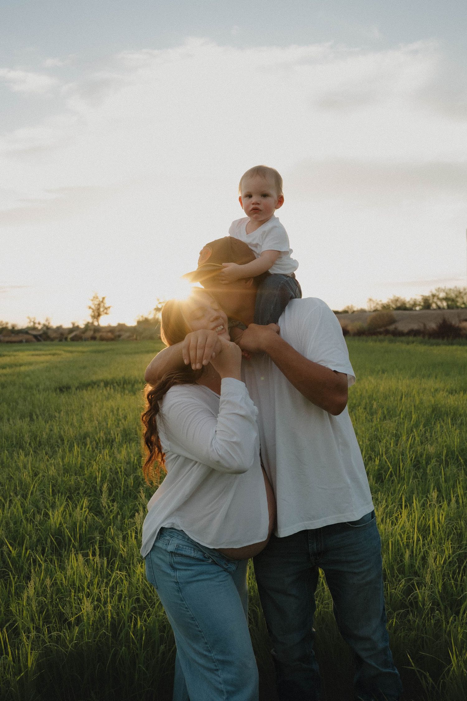 Family of three at sunset in green field, parents in white shirts holding baby on shoulders, golden hour glow.