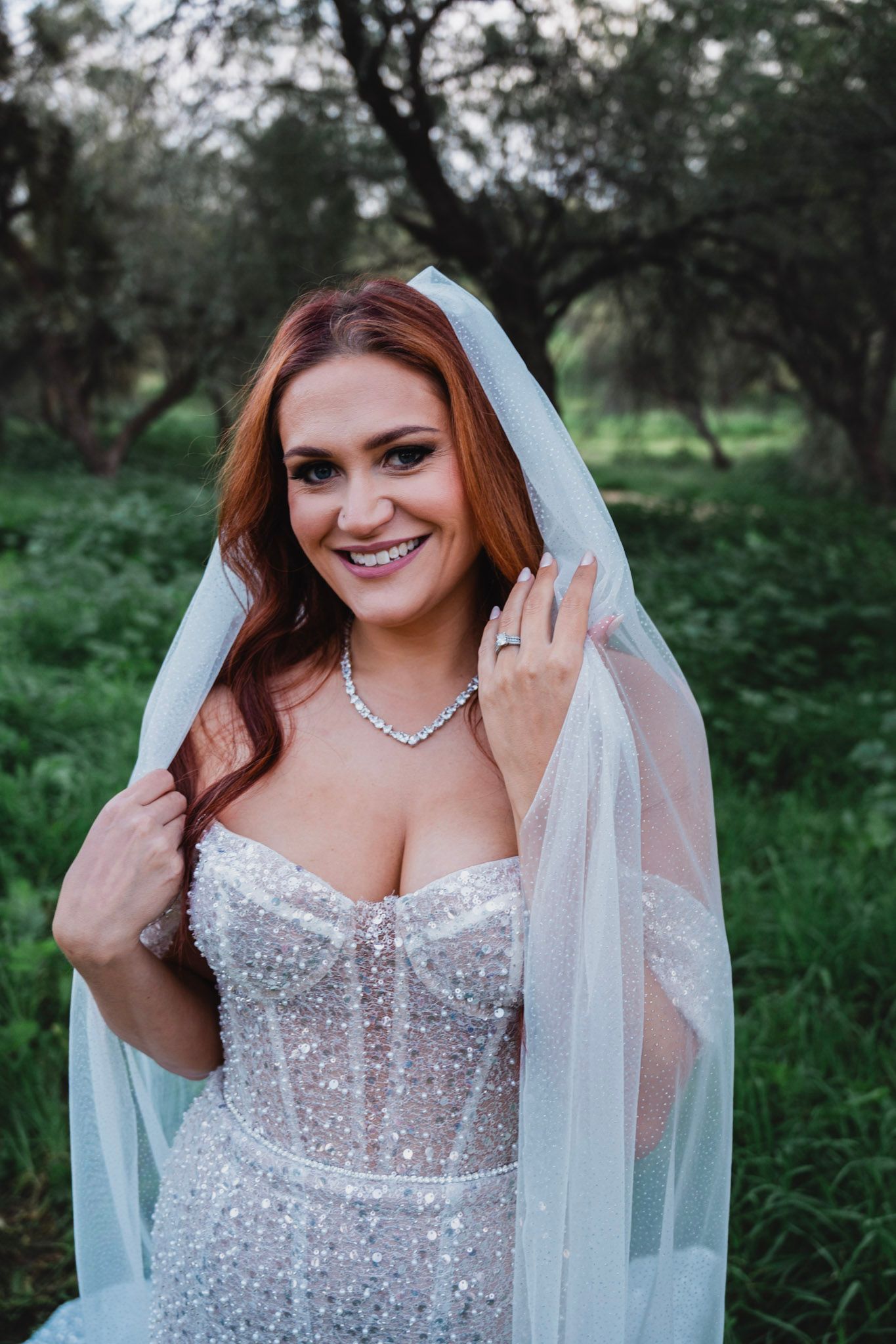 A smiling red-haired bride in a beaded gown lifts her veil in a lush green outdoor setting.