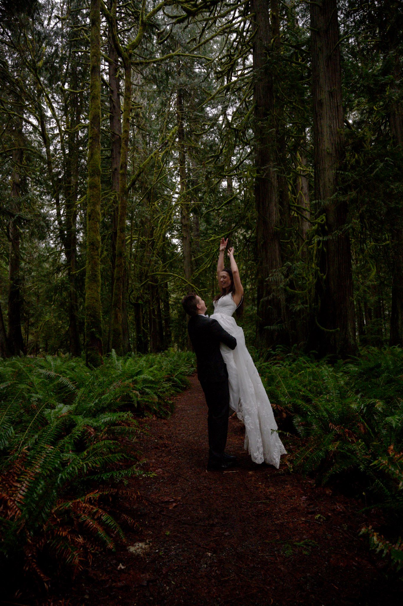 Bride and groom share a romantic moment on a forest path surrounded by towering trees and lush ferns.