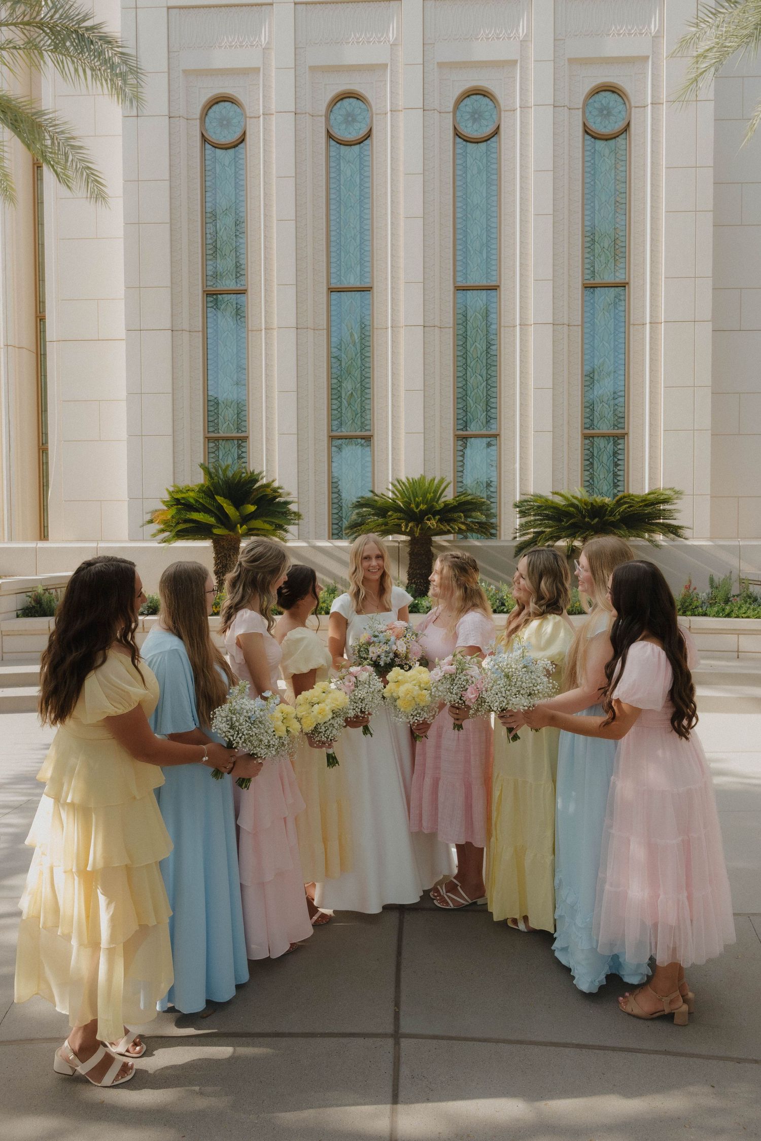 Bride surrounded by bridesmaids in pastel dresses holding white bouquets outside a white temple.
