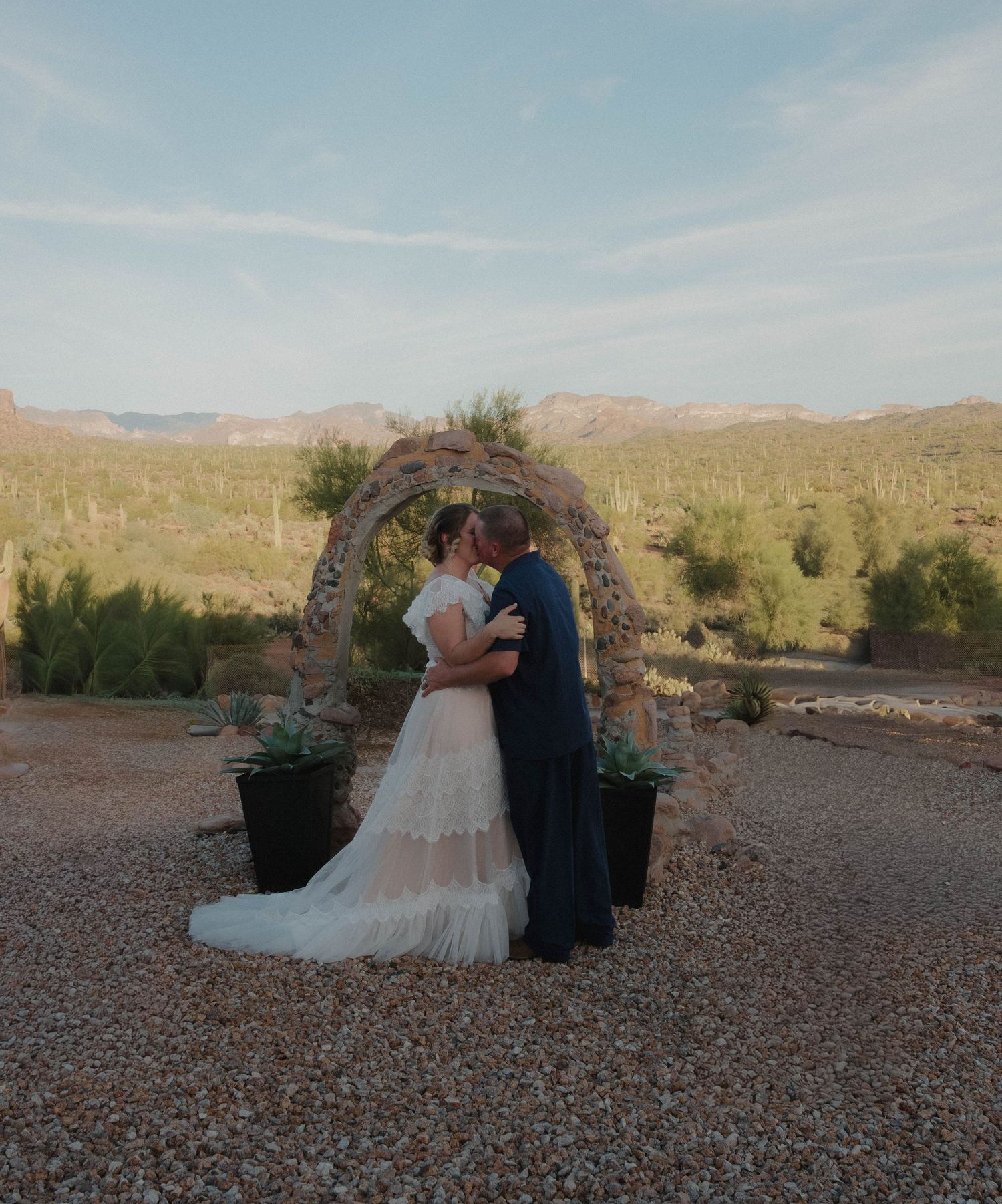 A couple shares their first kiss under a rustic arch in the Arizona desert at sunset during their outdoor wedding ceremony.
