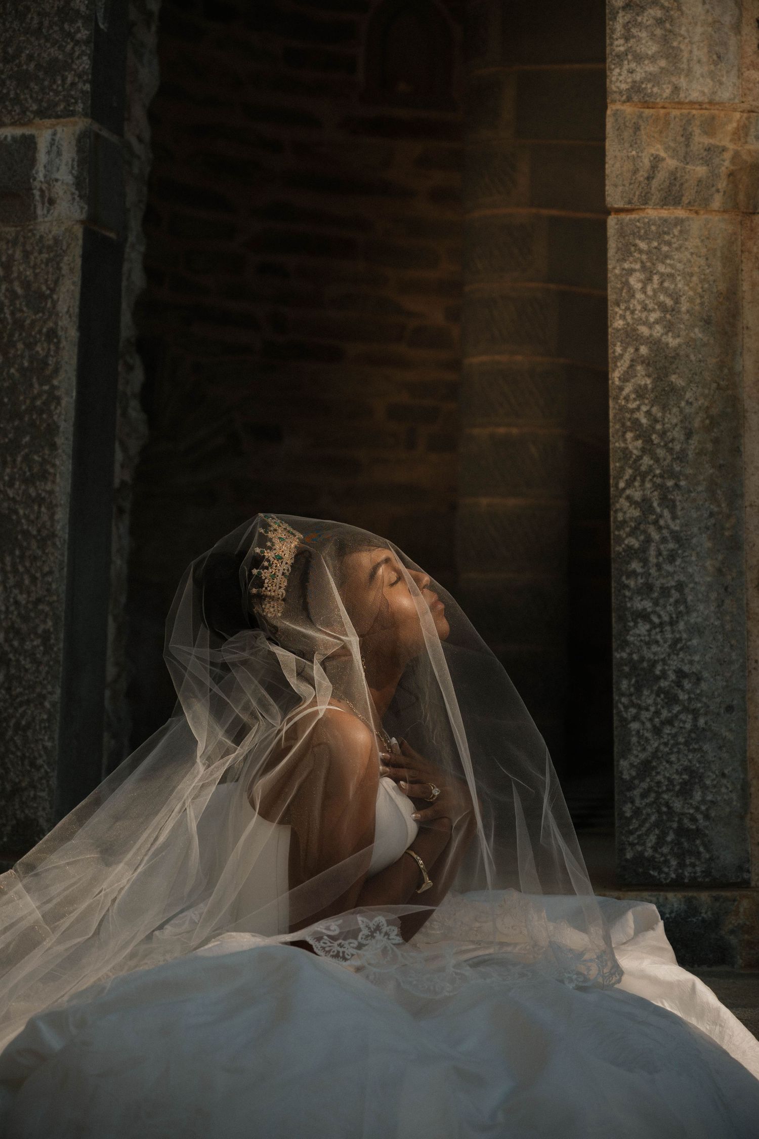 Bride in white gown and veil with pearl headpiece poses gracefully against stone architecture in dramatic lighting.