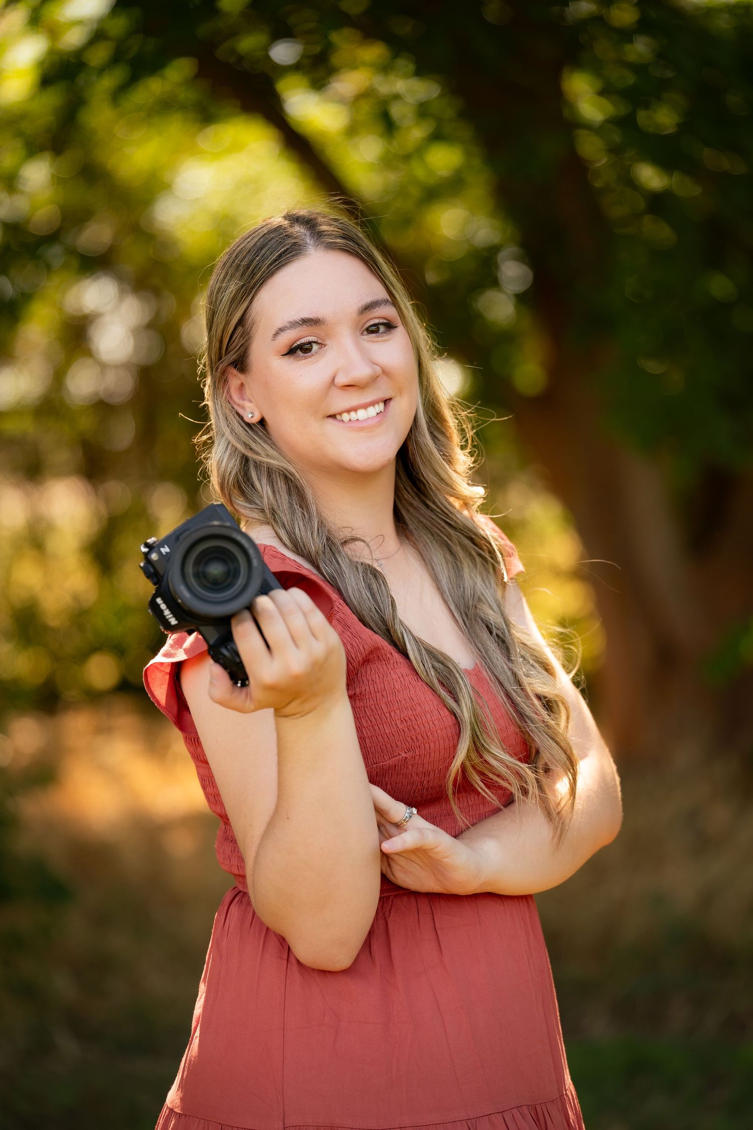 Smiling young woman in coral dress holding a Canon camera outdoors in warm sunlight.