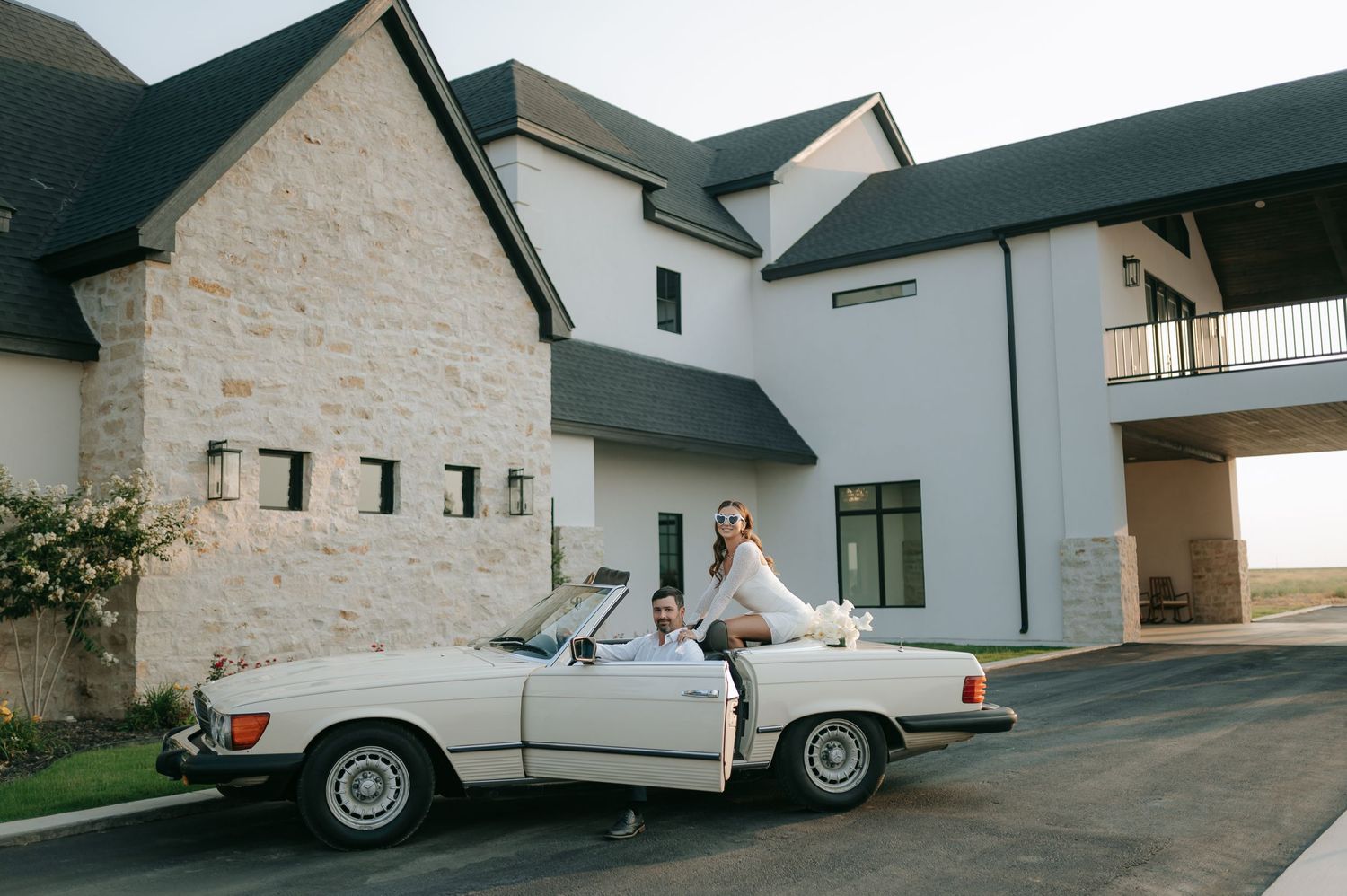 A bride and groom pose with a classic white Mercedes convertible outside a modern stone building.