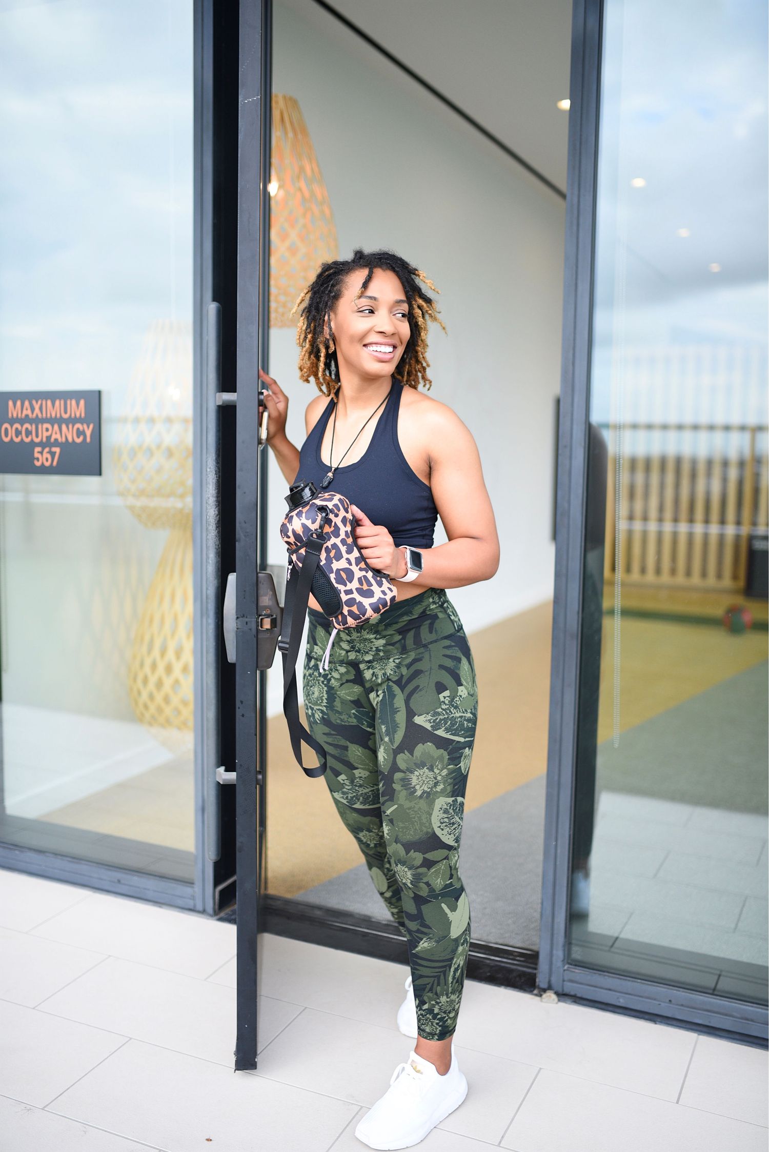Fit woman in camo leggings and black sports top smiling while holding door open at modern gym entrance.