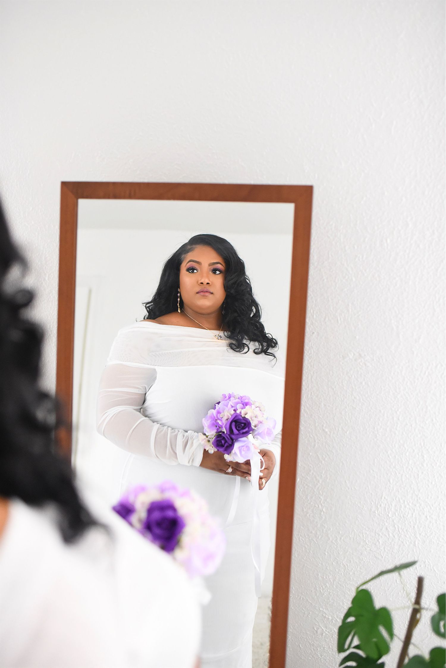 Woman in a white long-sleeve off-shoulder gown holding purple bouquet reflected in a full-length mirror.