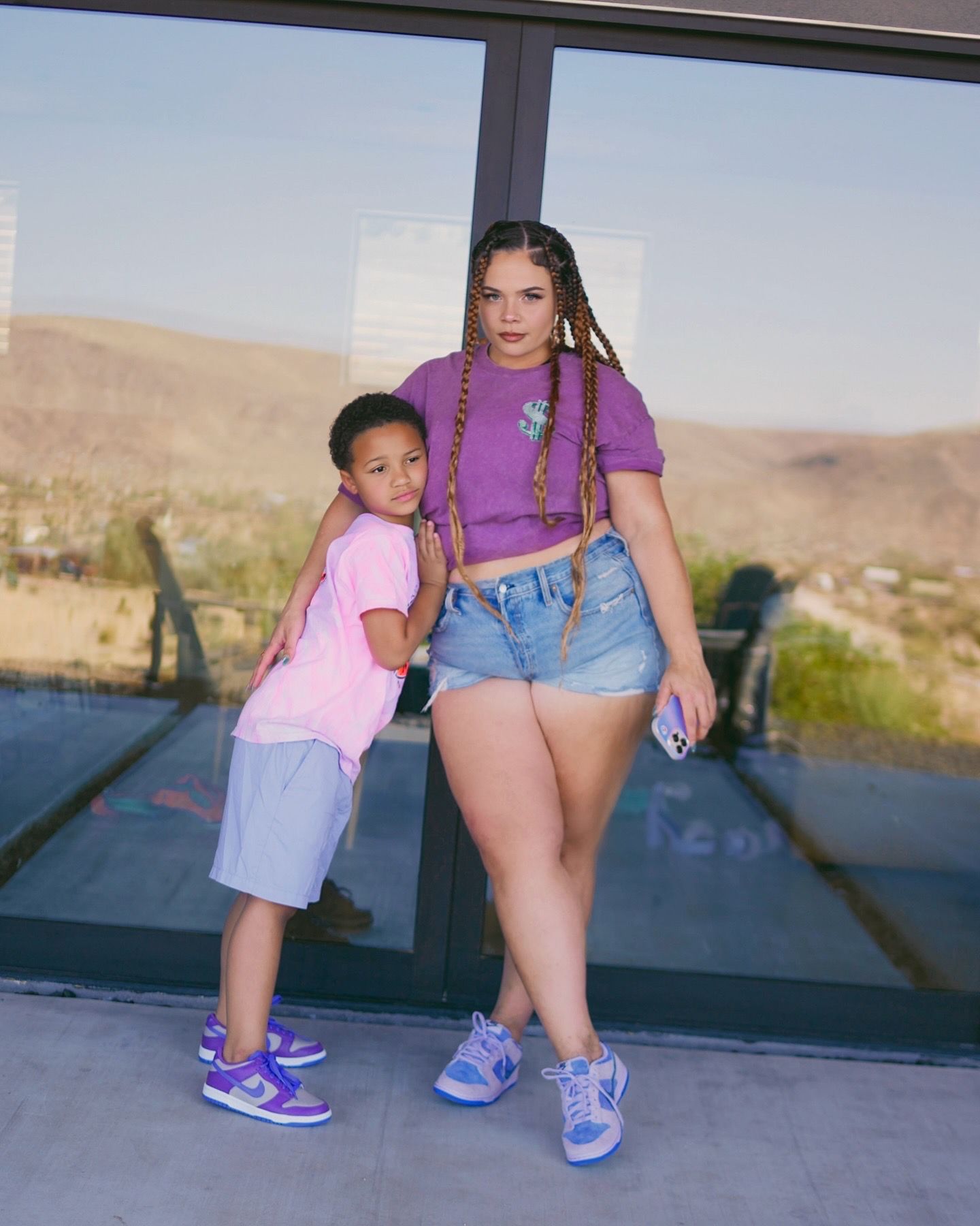 Woman in purple top and denim shorts standing with young child in pink outfit against glass window outdoors.