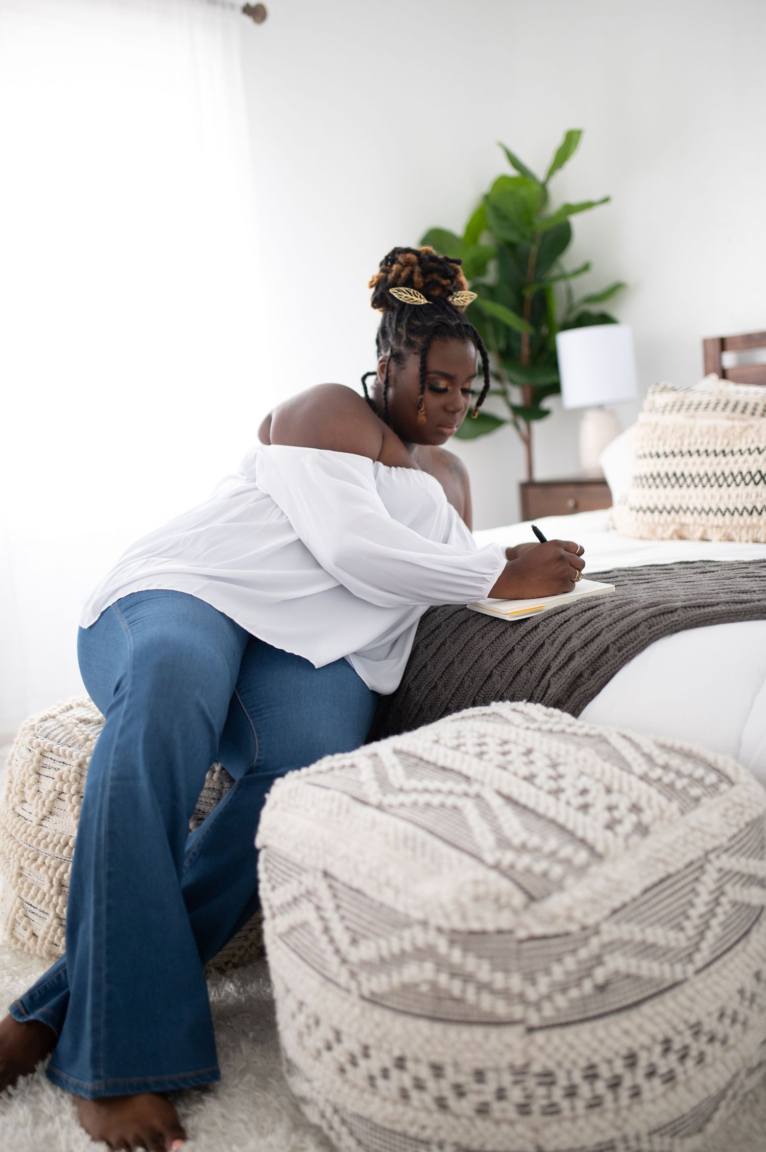 Woman in white off-shoulder blouse and flared jeans writes in journal on couch.