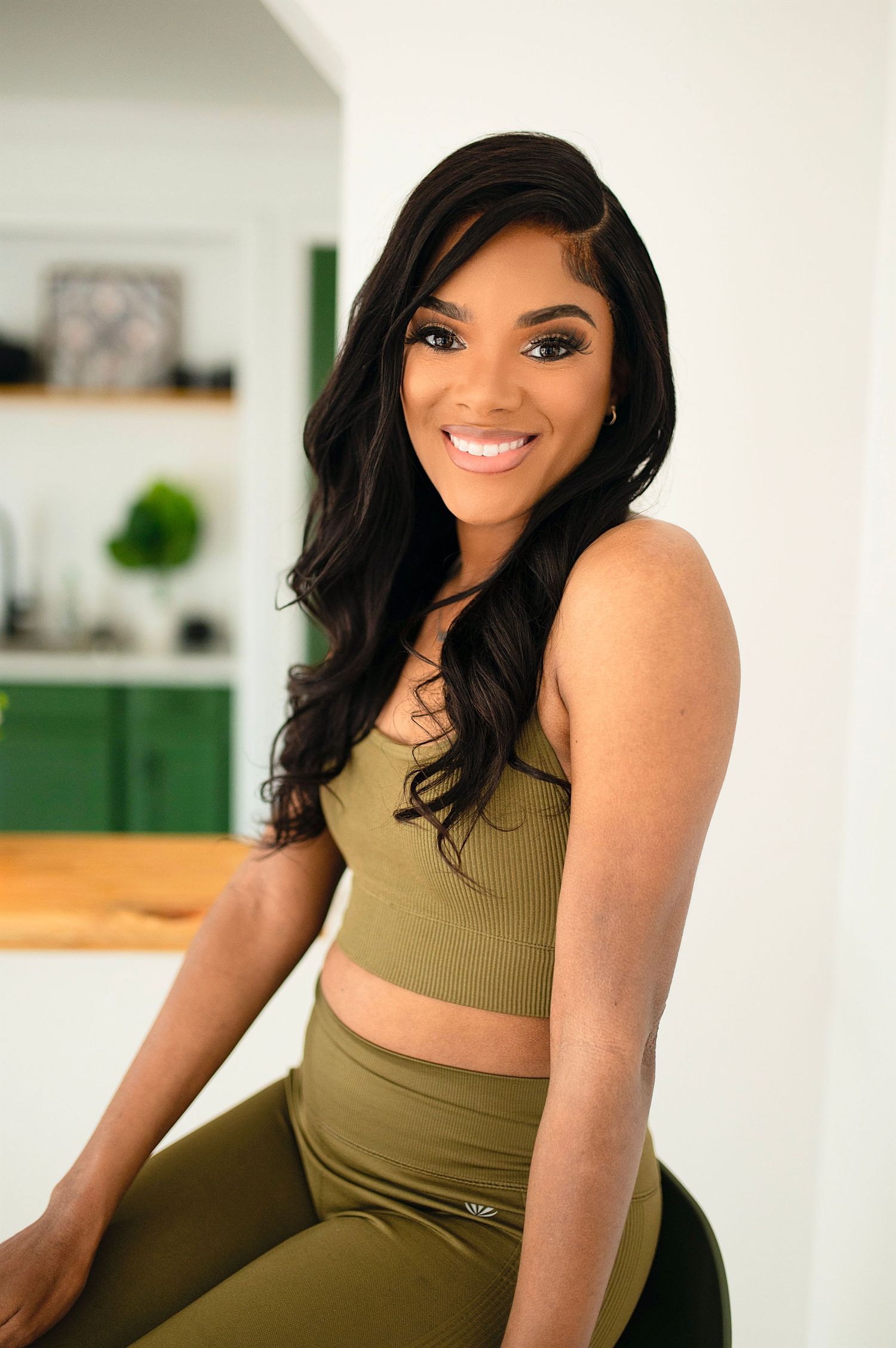 Smiling woman with long dark hair wearing olive green crop top and leggings indoors.