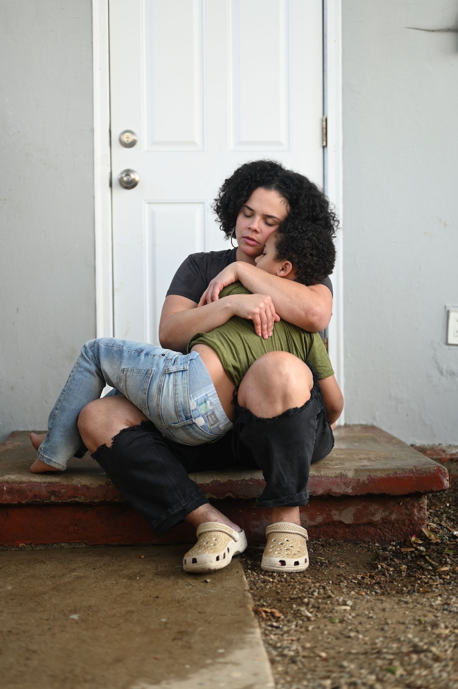 Black mother tenderly embracing young son on front porch steps in emotional moment.
