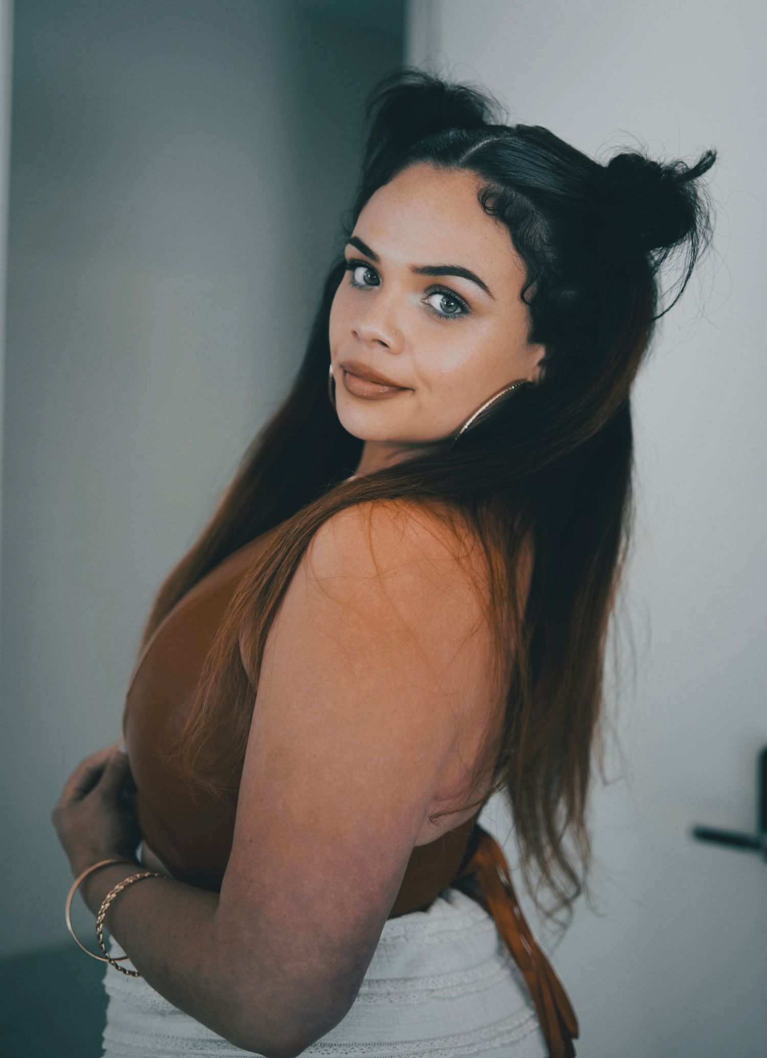 Stylish young woman with long dark hair and blue eyes poses confidently in a brown top and white skirt.