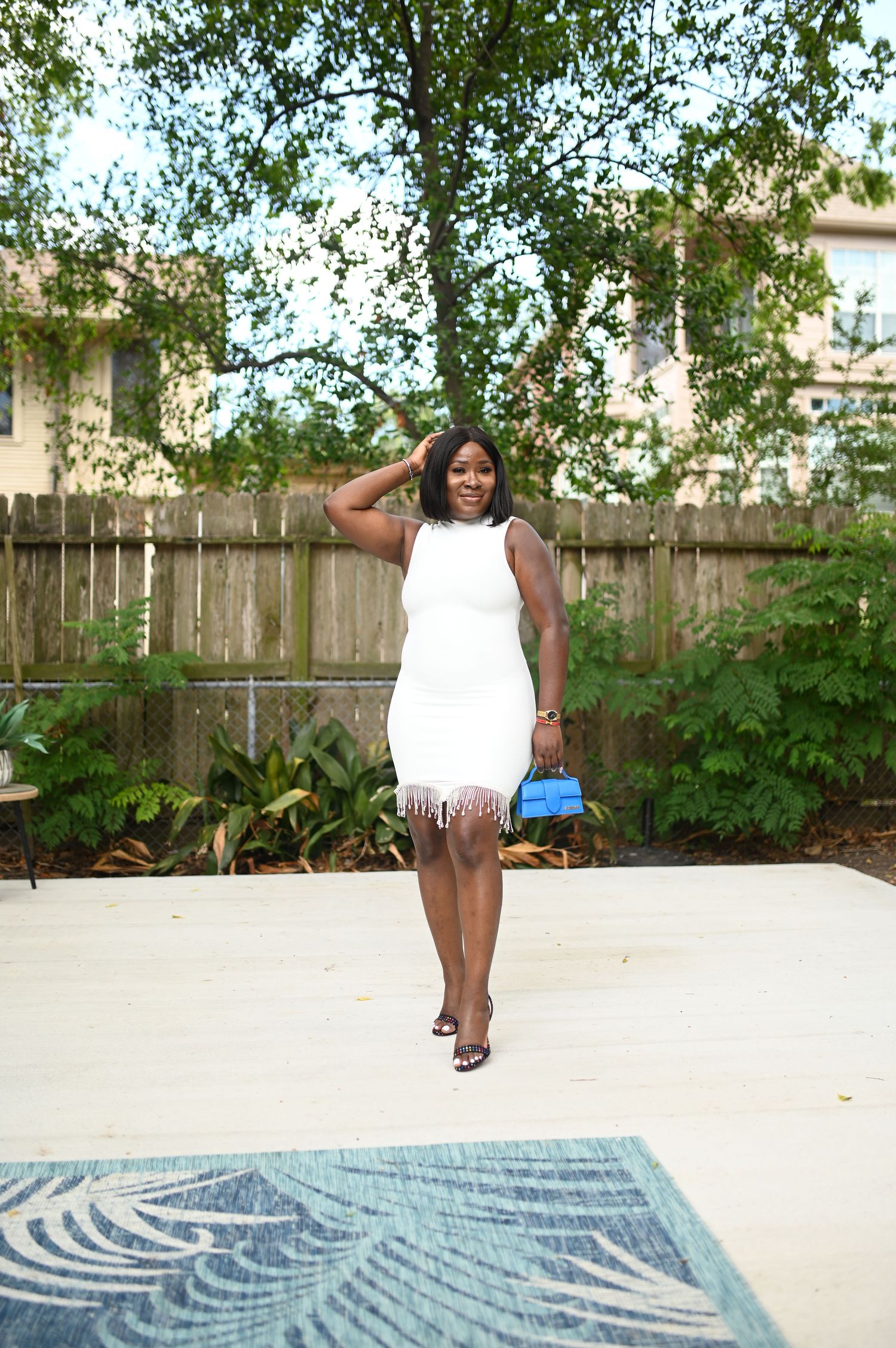 Woman in white fringed mini dress holds blue clutch while posing in outdoor backyard patio setting.