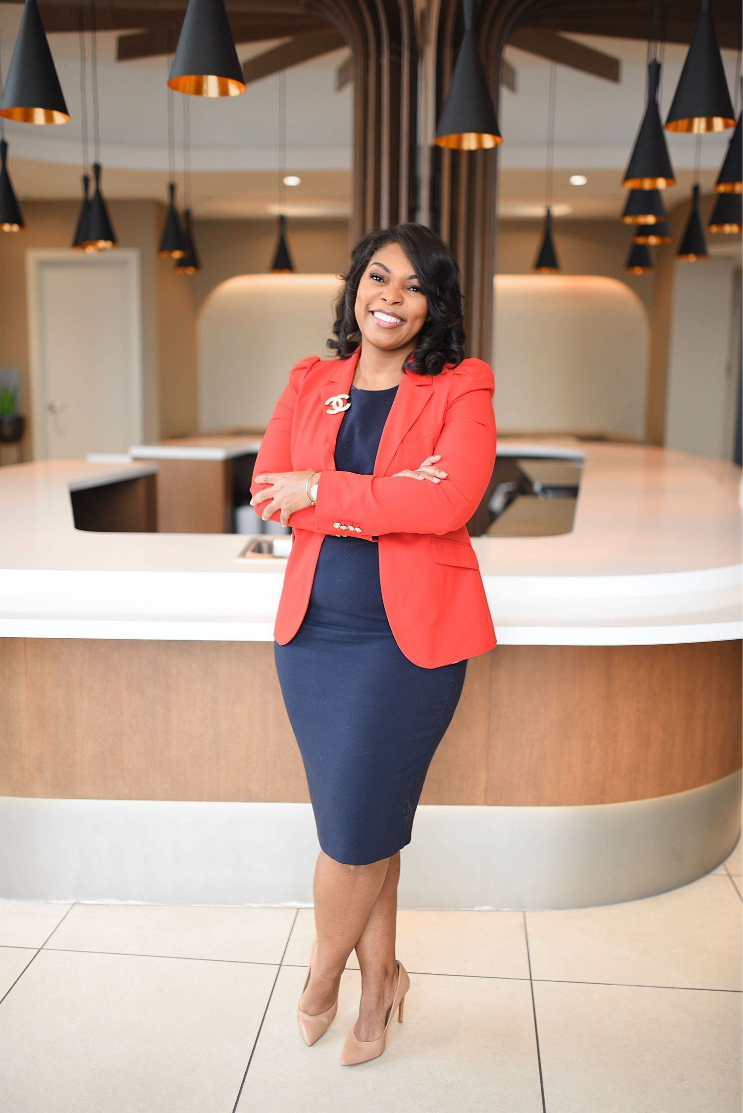 Woman in red blazer and navy dress posing confidently with arms crossed in stylish modern lobby.