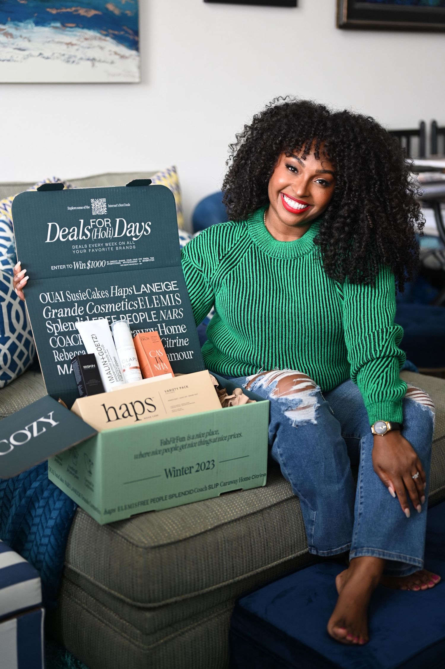 Smiling woman in green sweater and jeans holding a Haps subscription box on a couch.