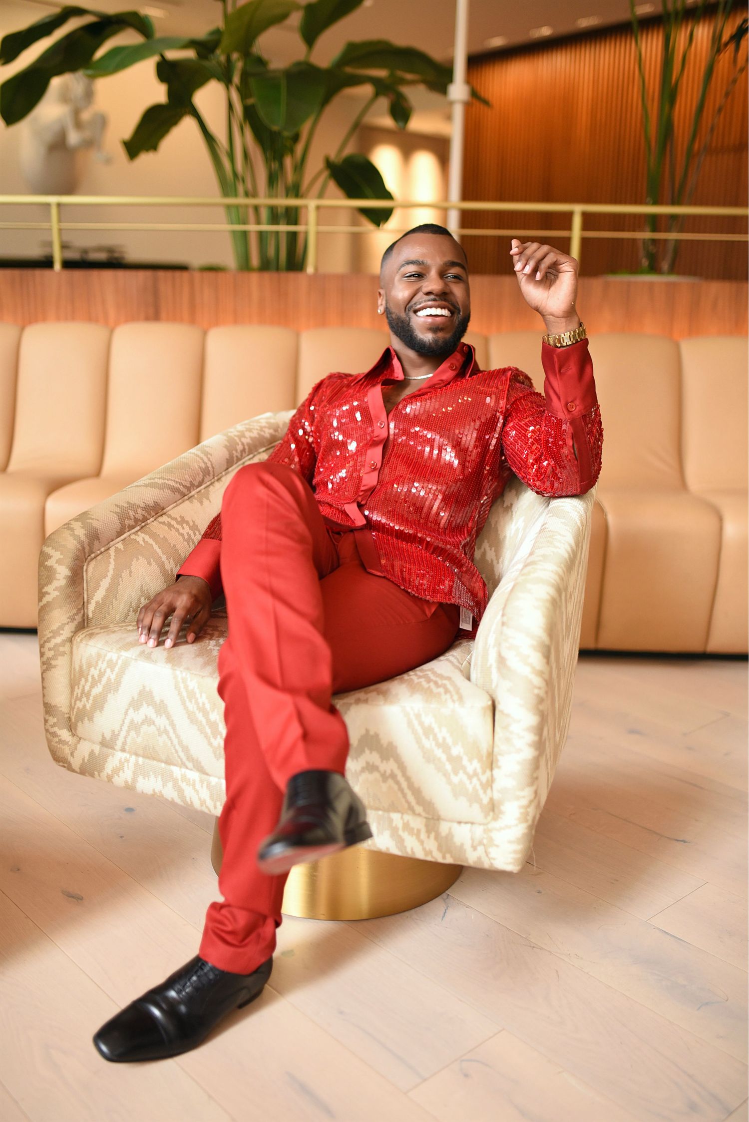 Man in red sequin shirt and pants laughing joyfully while seated in a gold velvet chair.