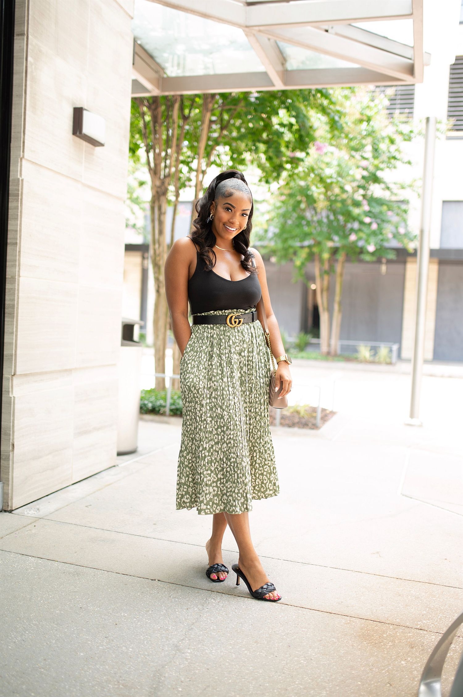 Young woman in floral midi skirt, black bodysuit, and Gucci belt poses outdoors near modern building.