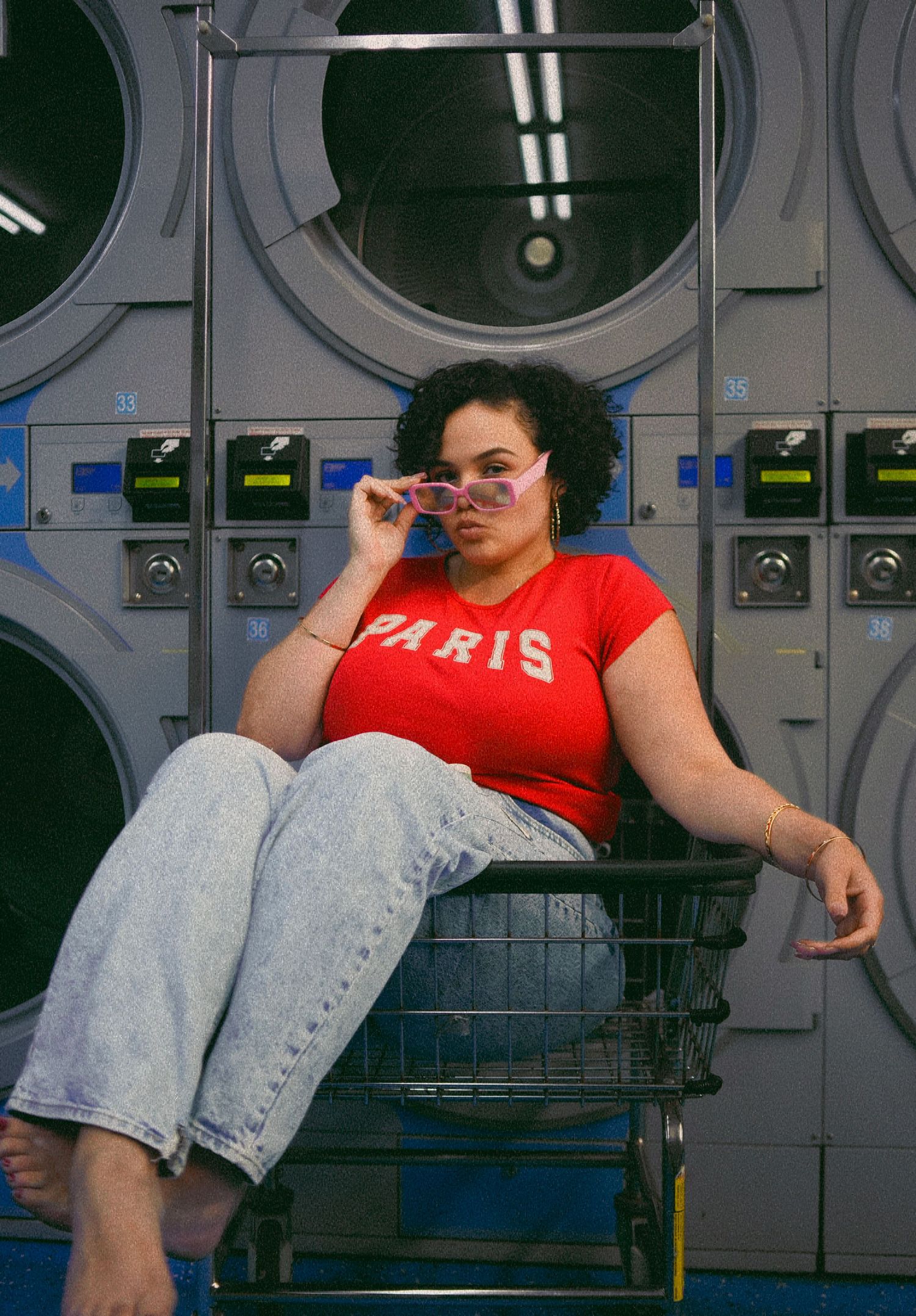 Curly-haired woman in red Paris top seated in laundry basket at laundromat.