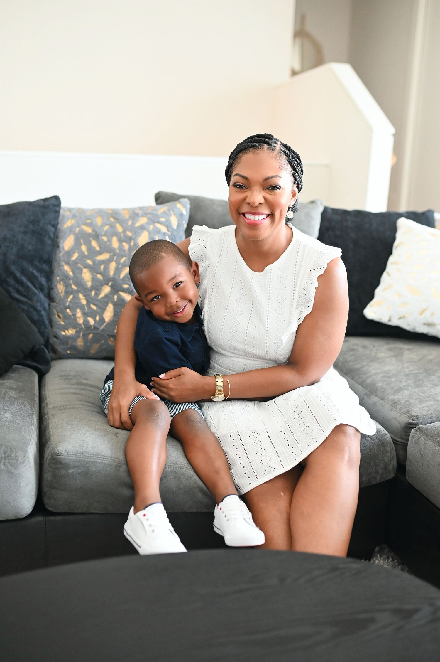 A happy Black mother and young son sit together on a gray couch indoors.